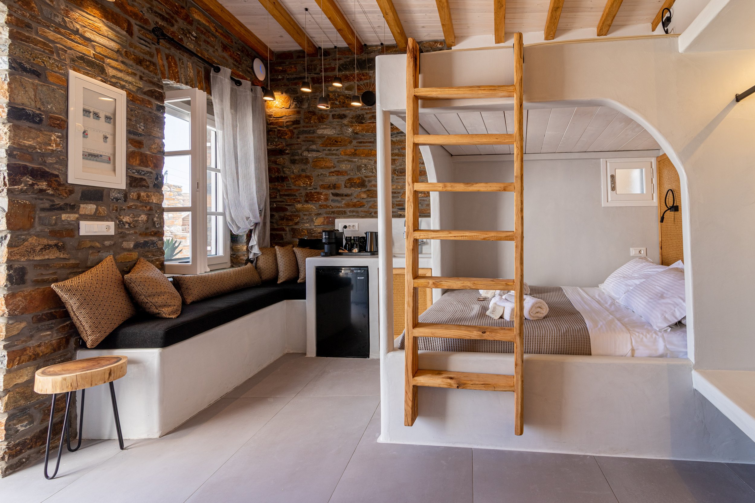 Interior of a cozy studio apartment with a loft bed, a sitting area with cushions, a window with curtains, a small refrigerator, and a kitchenette.