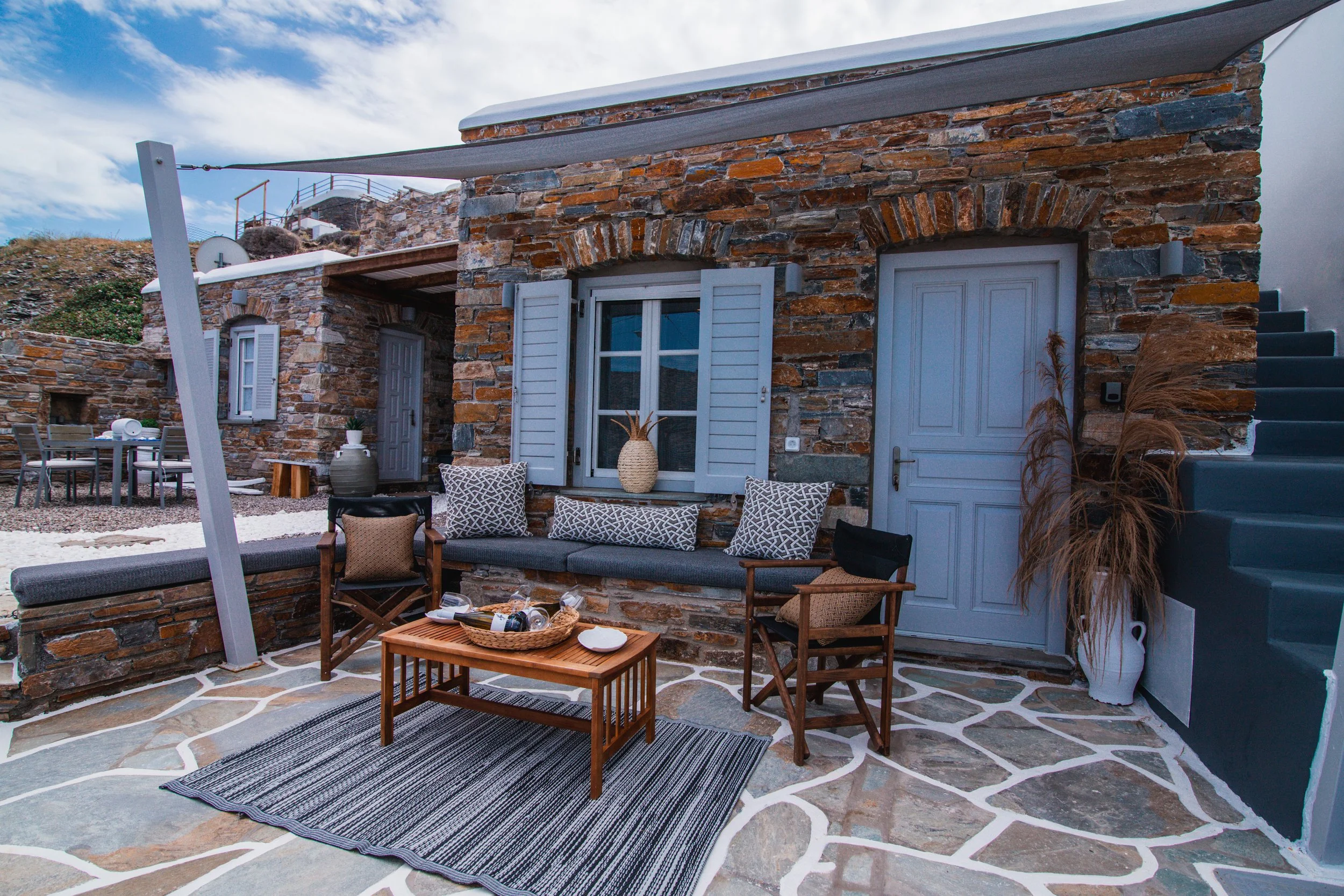 Outdoor patio with stone walls, a cushioned bench with patterned pillows, a coffee table with decorative items, and wooden chairs, in front of a house with blue shutters and a door, under a canopy.