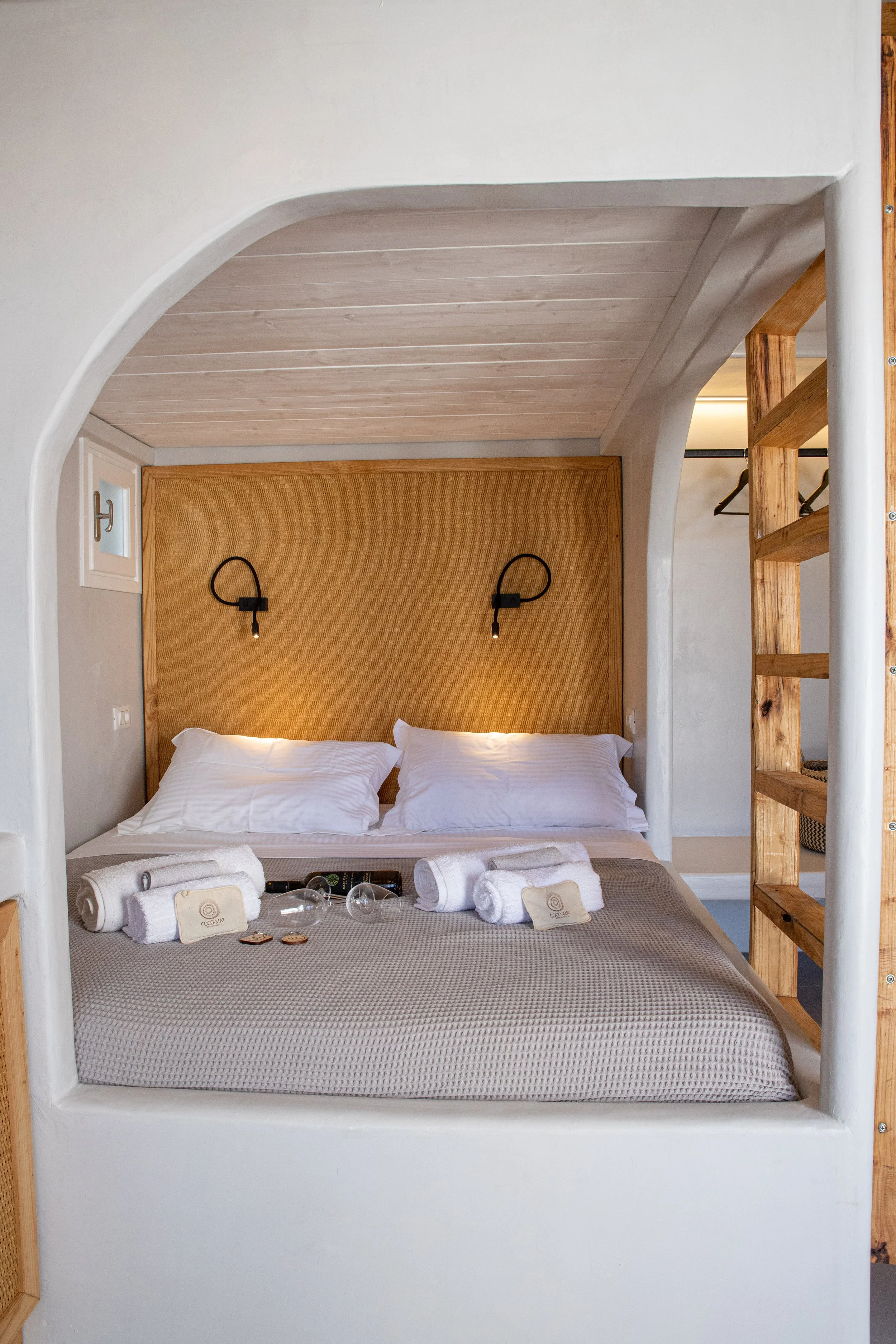 A cozy hotel room with a neatly made bed, white bedding, and four rolled towels on top. The bed has a wooden panel headboard with two black wall-mounted bedside lamps. The room features white walls, wooden accents, and a small built-in shelf. The ove