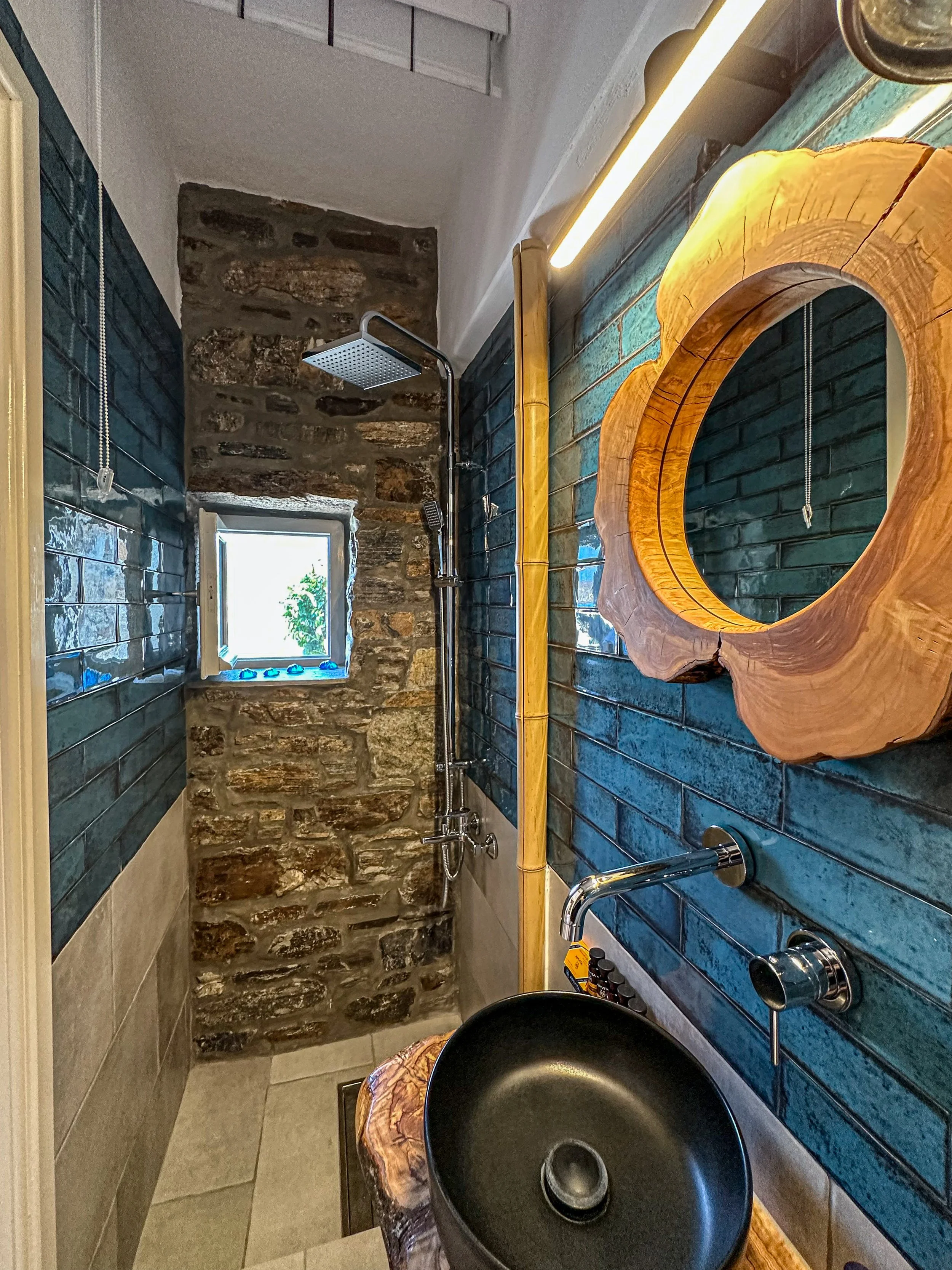 Small rustic bathroom with exposed stone wall at end of shower and a small window. Dark blue tiled wall with a natural wood framed mirror and sink. Shower has a rainfall head and handheld shower. Wooden accents and modern lighting.