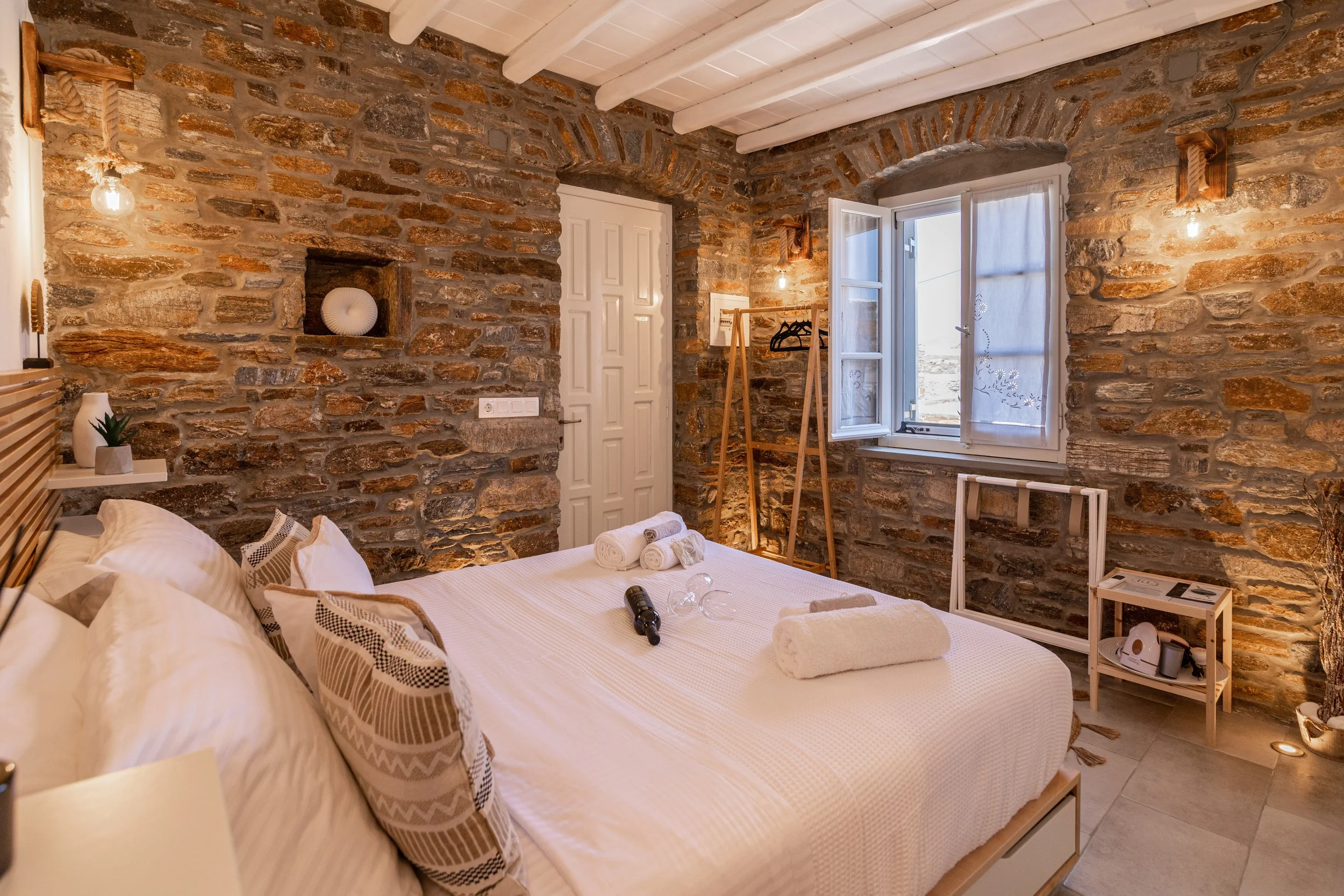 A cozy bedroom with a rustic stone wall, white bed with decorative pillows and rolled towels, an open window with sheer curtains, and warm lighting fixtures.