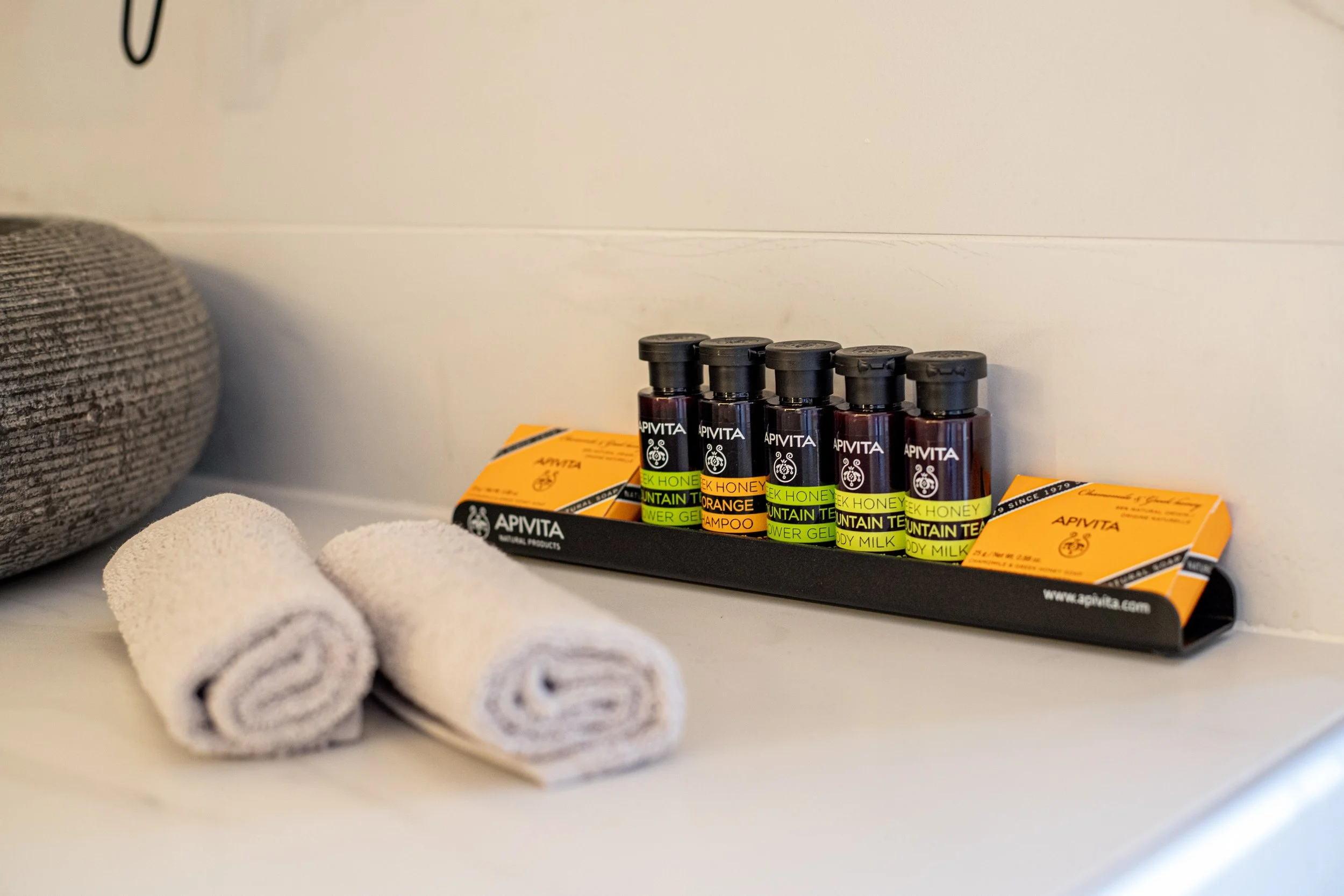 A set of small bottles of APIVITA herbal products, including body milk, body gel, shampoo, and hair mask, arranged on a black tray. Two rolled white towels are placed in front of the tray, with a textured gray cushion to the left.