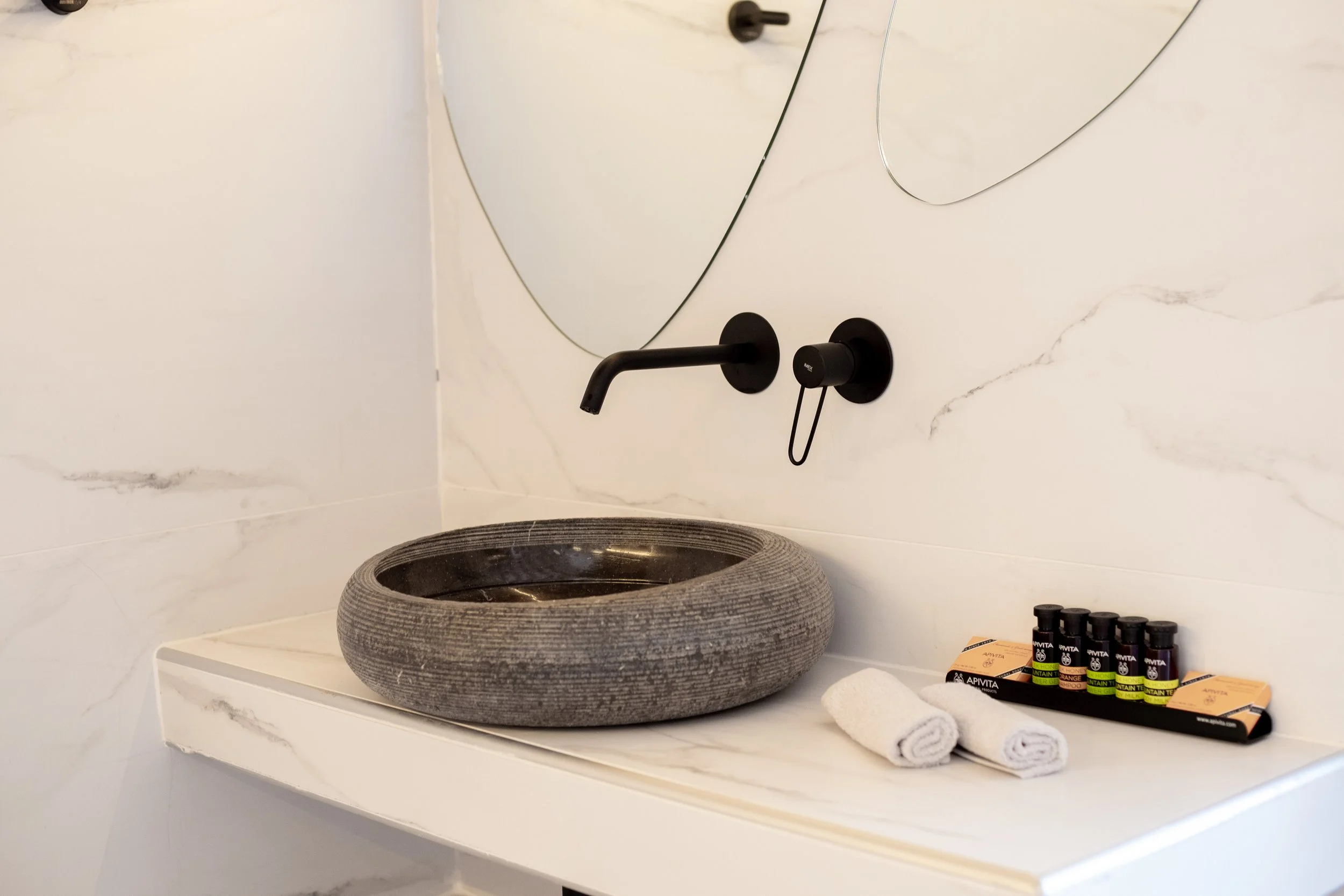 Bathroom vanity with a gray stone vessel sink, black wall-mounted faucet, bottles of essential oils, and two rolled white towels in front of a large marble wall mirror.