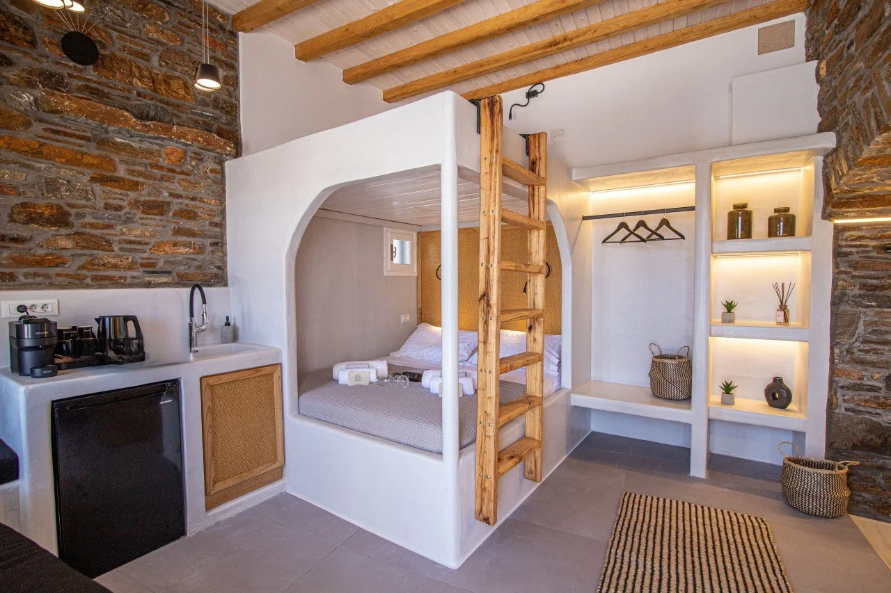 A compact studio with a lofted bed, wooden ladder, small kitchenette, and built-in shelves with decorative items, featuring exposed brick walls and modern lighting.