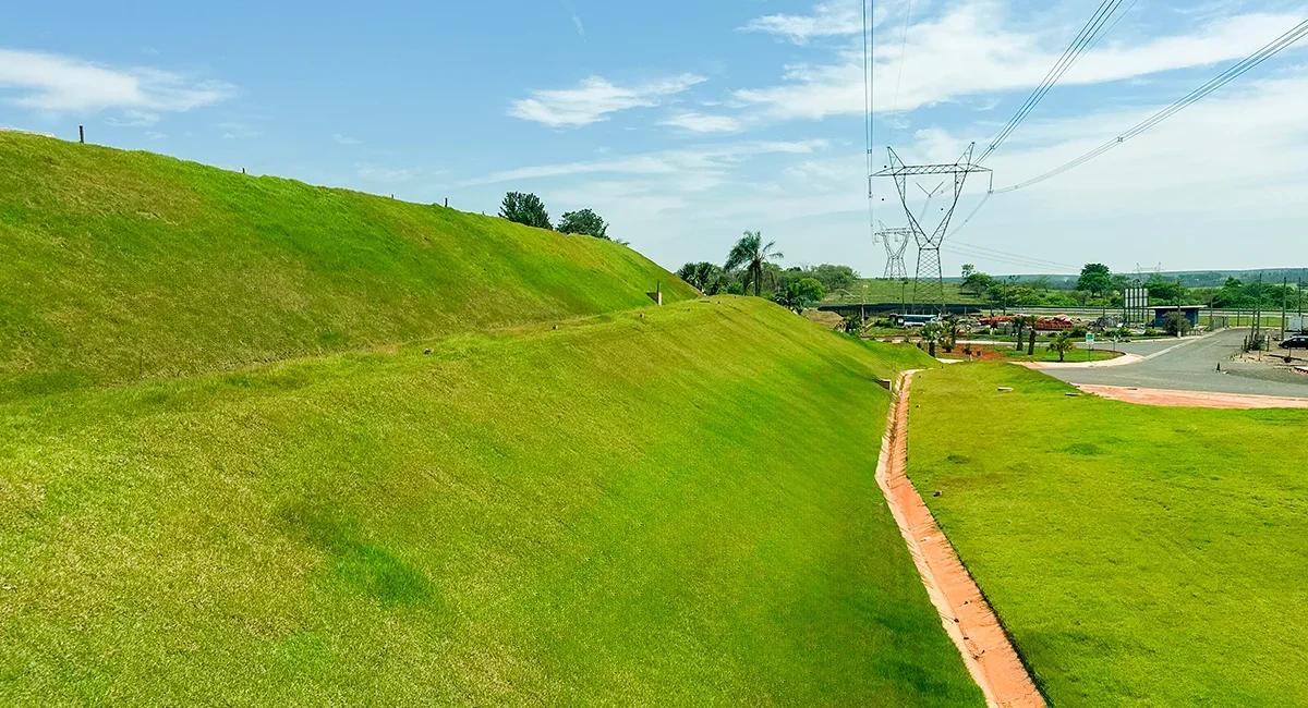 Área de grama verde com uma calçada de tijolos ao lado, arcs elétricos ao fundo e céu azul com algumas nuvens.