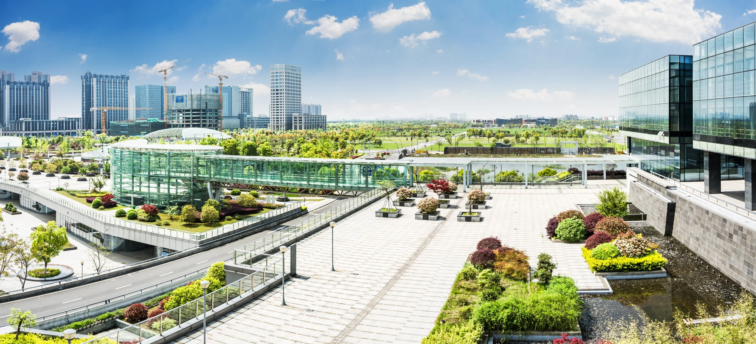 Vista de um prédio de escritórios com jardins e praça ao ar livre, com árvores, plantas e áreas de lazer, com cidade moderna ao fundo e céu azul com nuvens.