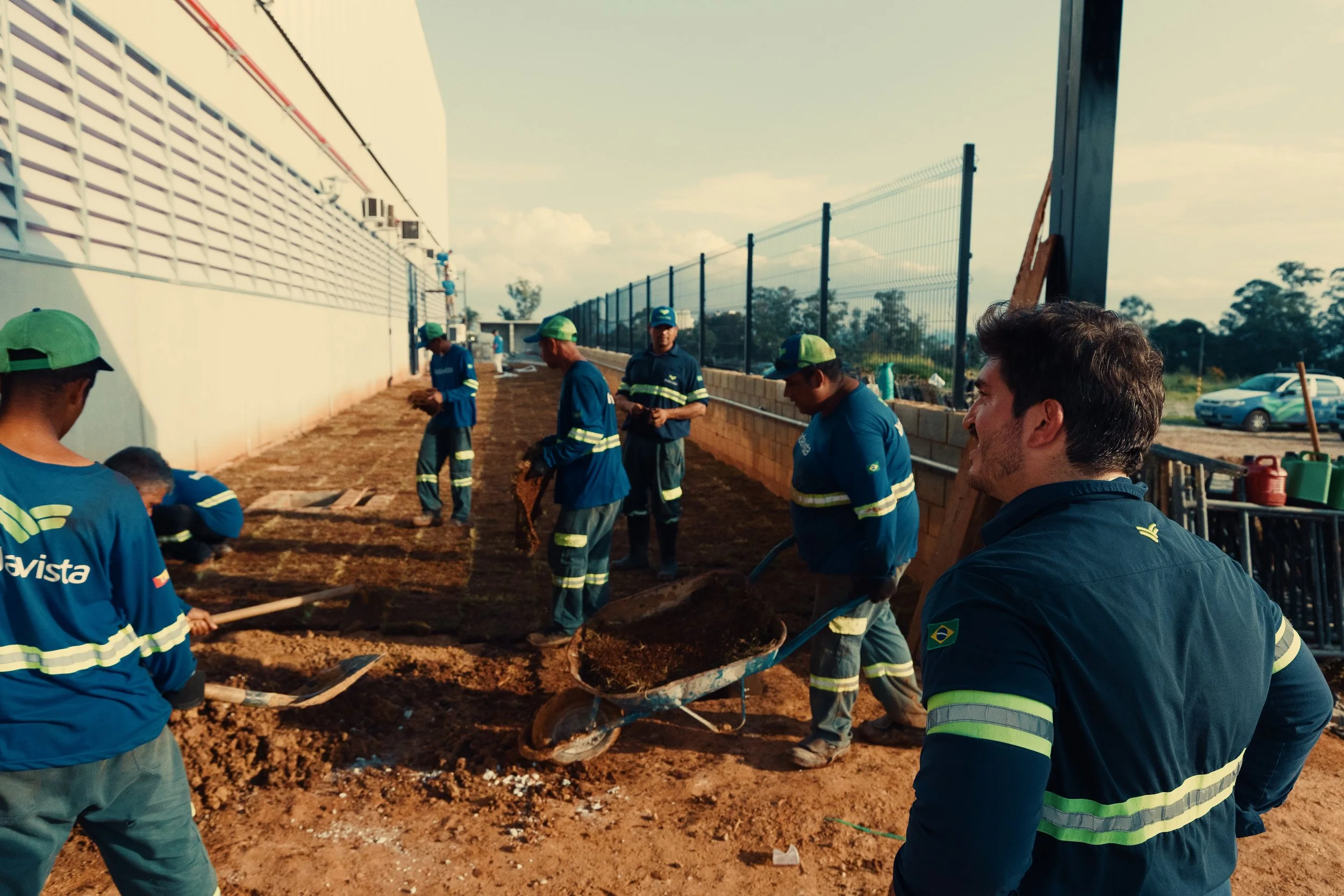 Vários trabalhadores usando uniformes azuis e bonés verdes trabalhando na construção de uma equipe de asfalto ao lado de um muro de concreto e uma cerca de metal, sob céu ensolarado.