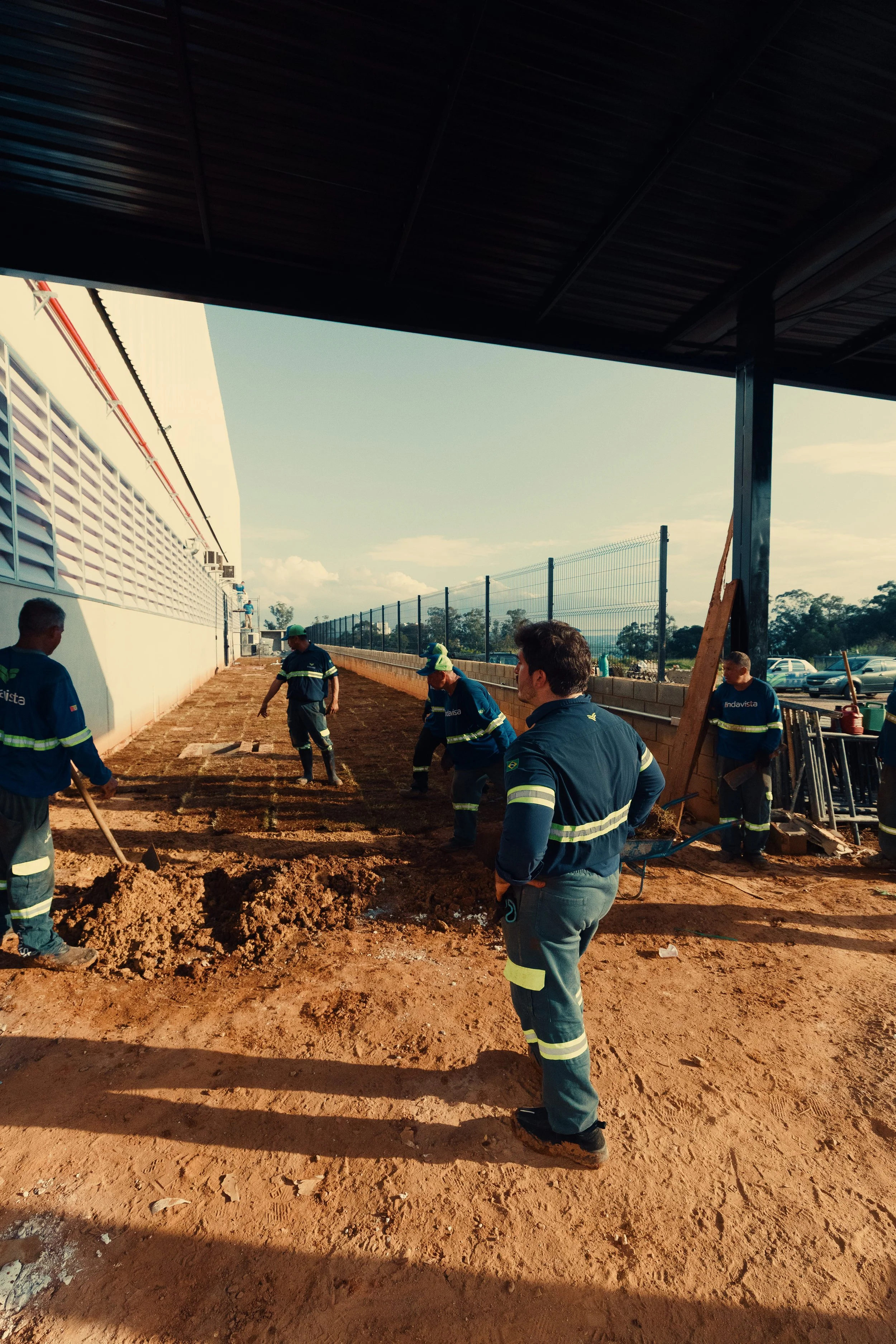 Homens trabalhando na construção de um piso de terra ao ar livre, com um galpão e uma cerca de metal ao fundo.
