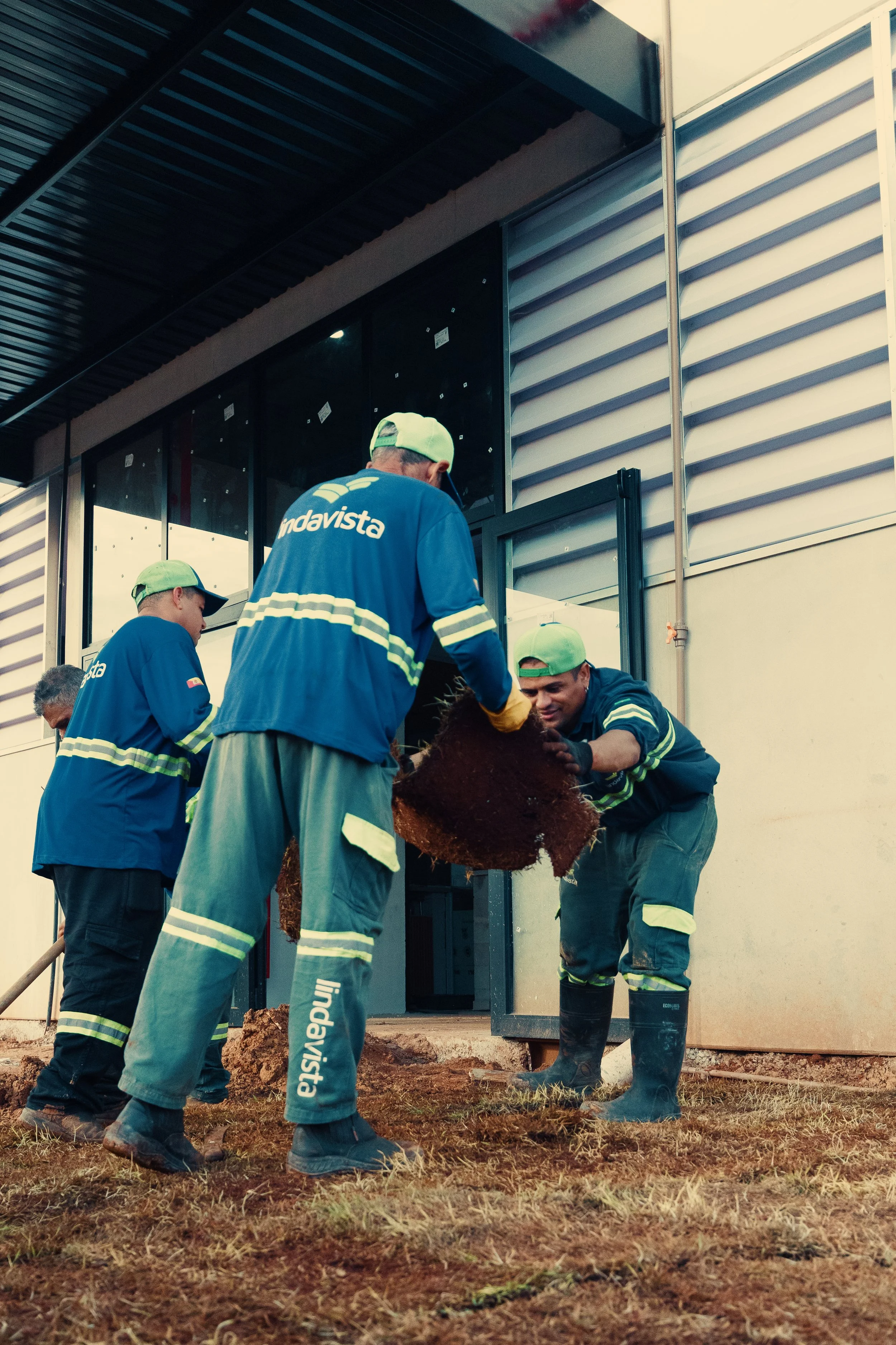 Trabalhadores mexendo na construção de uma estrutura, usando roupas de proteção e botas de borracha, ao lado de um edifício com paredes de metal e vidro.