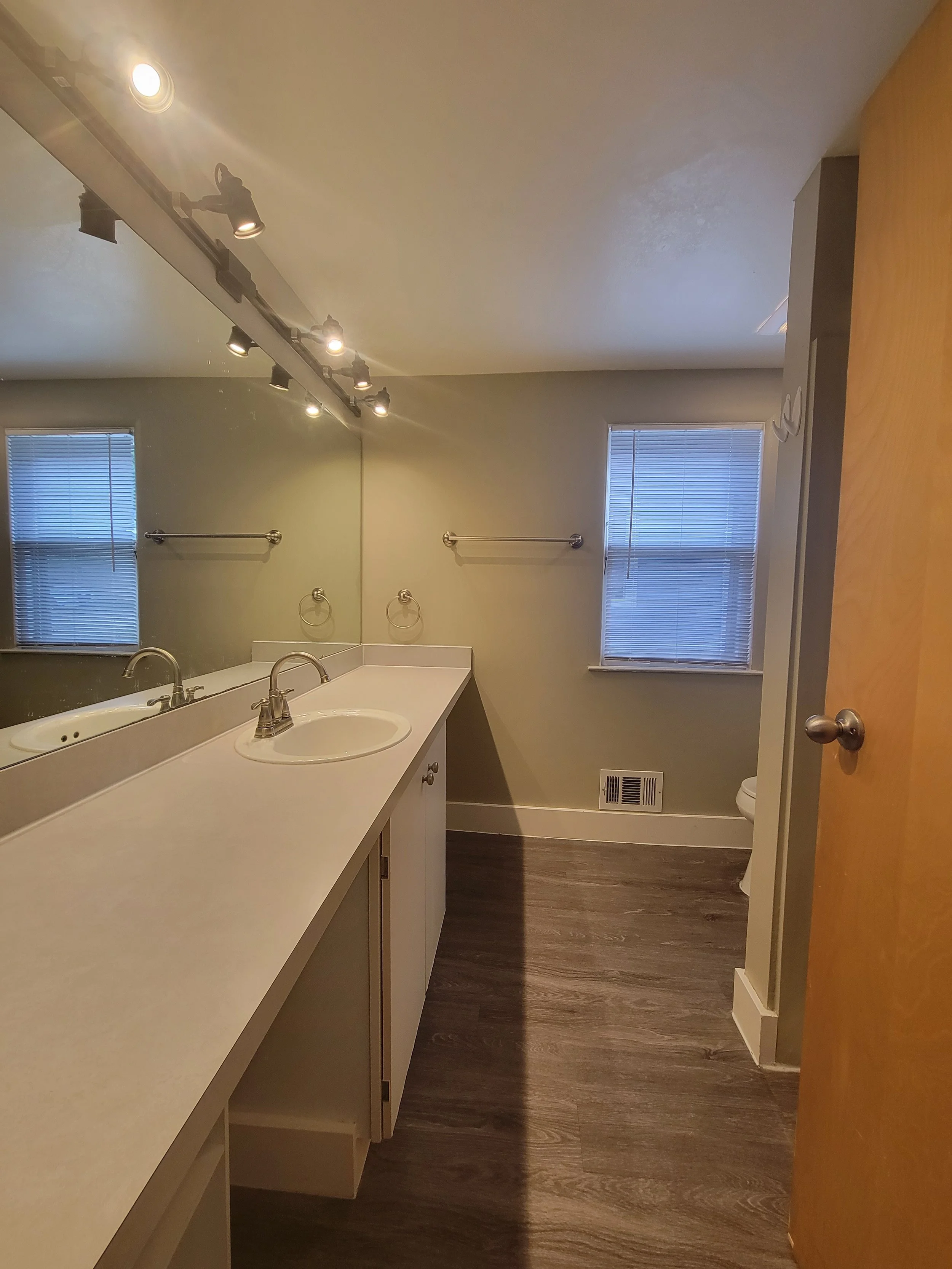 Modern Ann Arbor home with wood flooring, large mirror, sink with faucet, track lighting, towel racks, and small window with blinds.