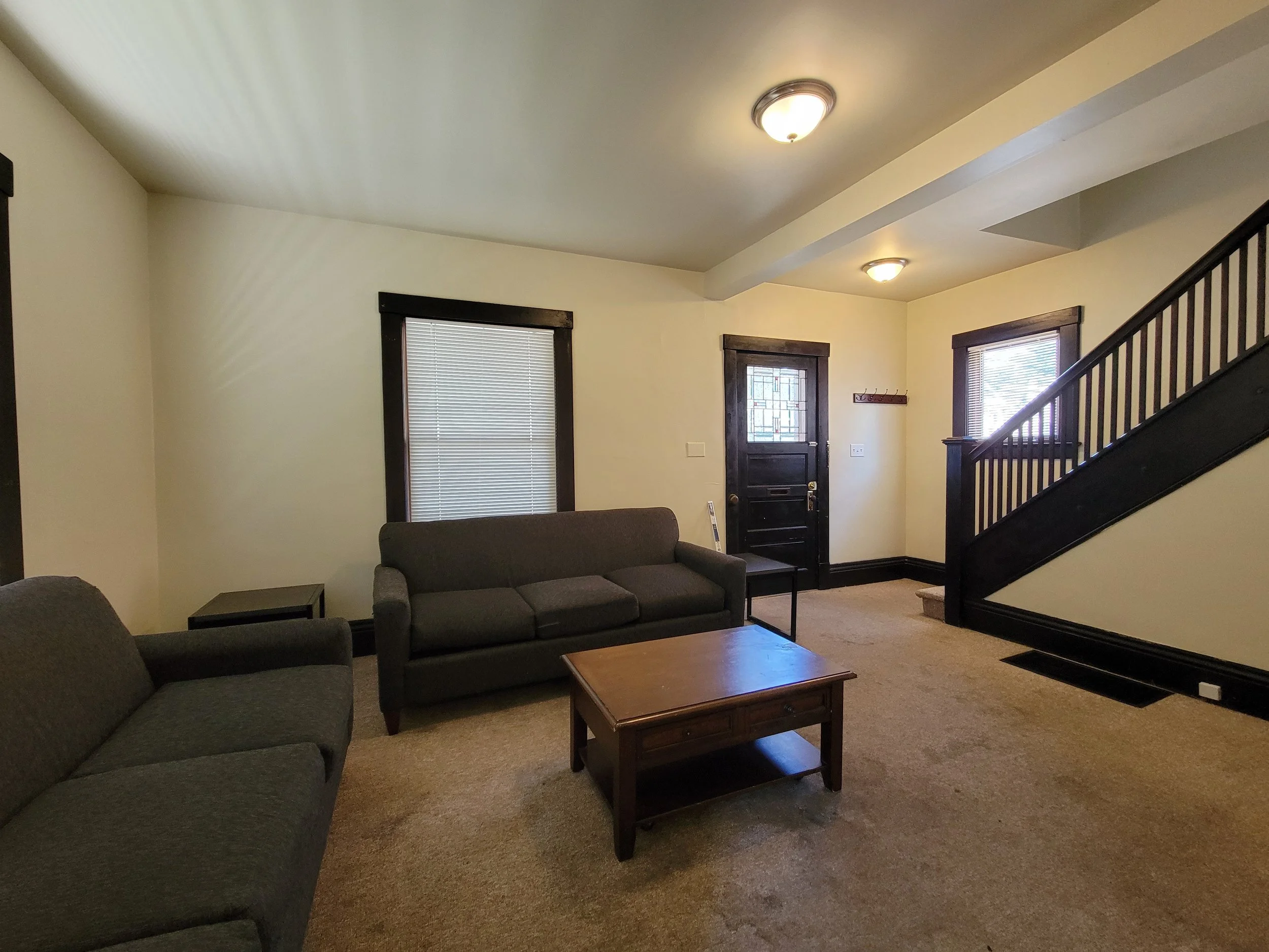 Living room home rental in Ann Arbor with two gray couches, a wooden coffee table, and a staircase. The room has beige walls with dark trim, a door with a decorative glass panel, and carpeted floor.