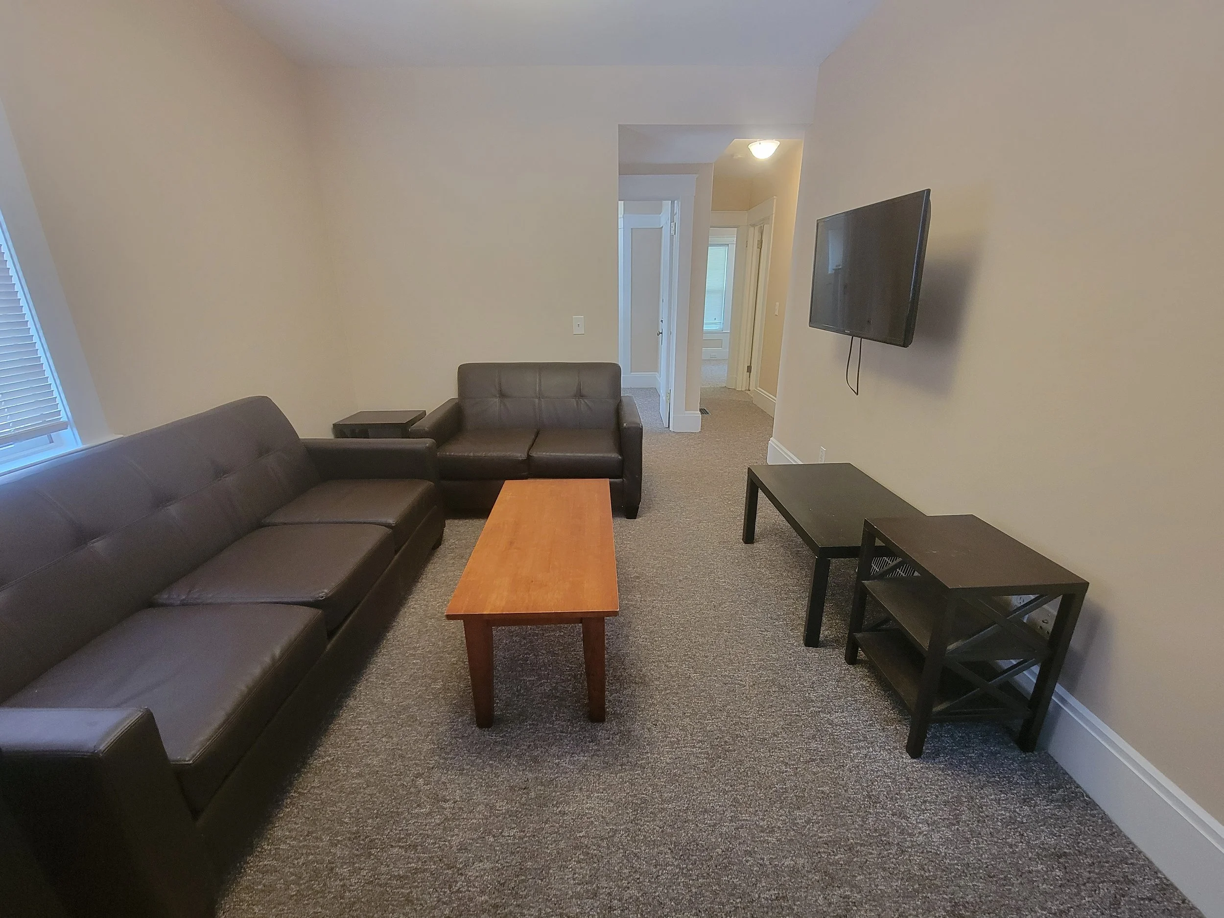 A home for rent with two dark leather sofas, wooden coffee table, flat-screen TV on the wall, and two side tables on a carpeted floor.