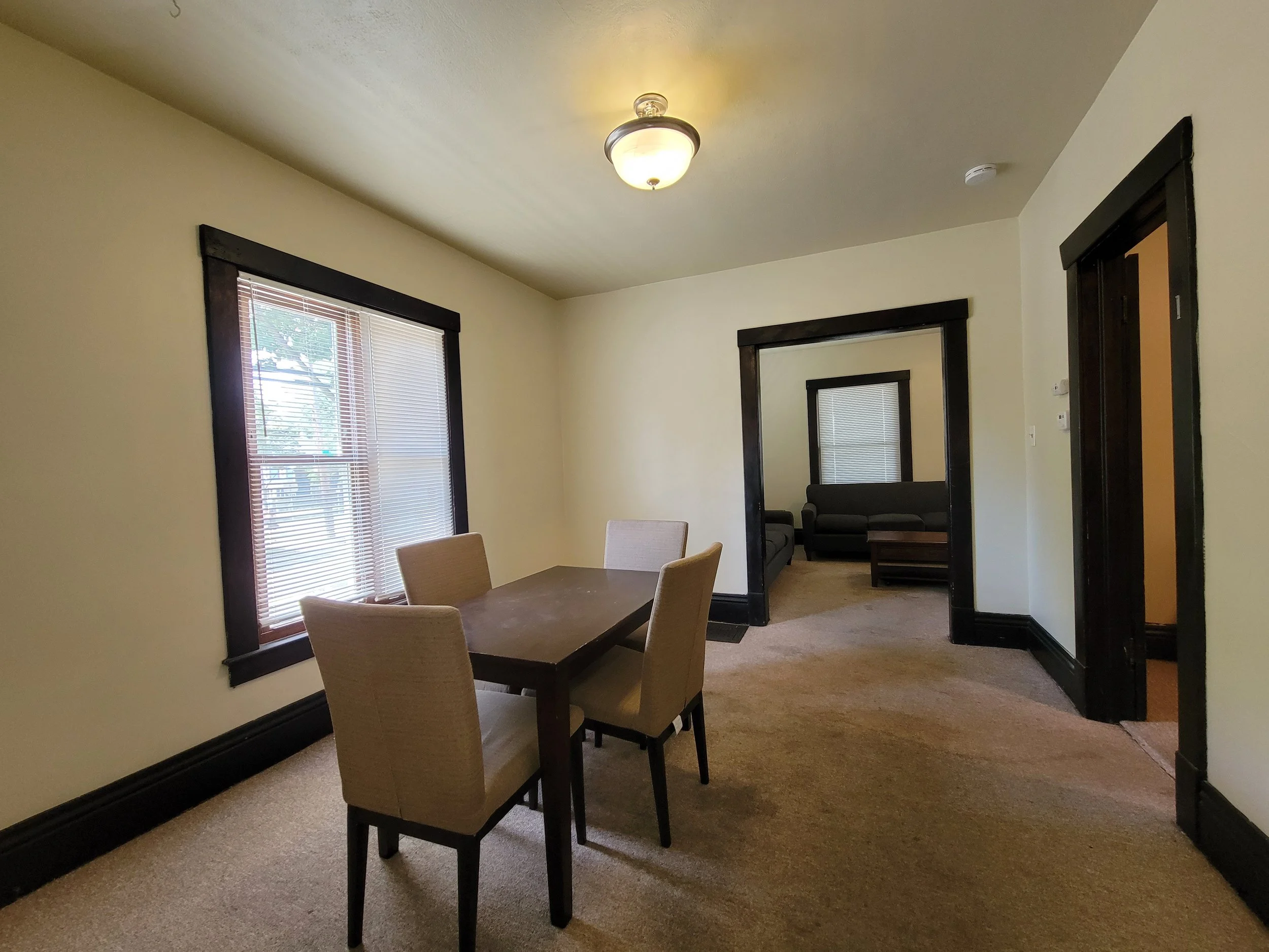 Rental property in Ann Arbor with a wooden table and chairs, beige walls, wall-mounted light fixture, window with blinds, and view into a living room with couches and a coffee table.