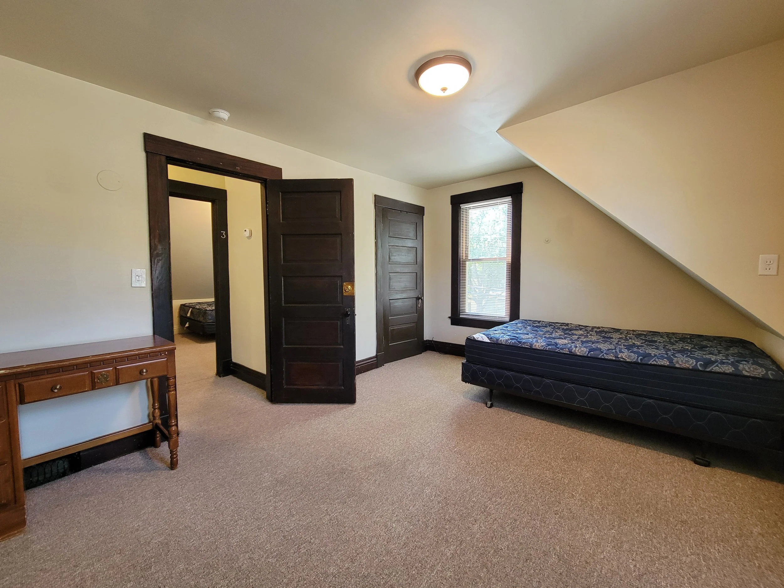Room interior with a twin bed and small wooden desk, beige walls, brown wooden trim, and carpeted floor in warm rental house in Ann Arbor.