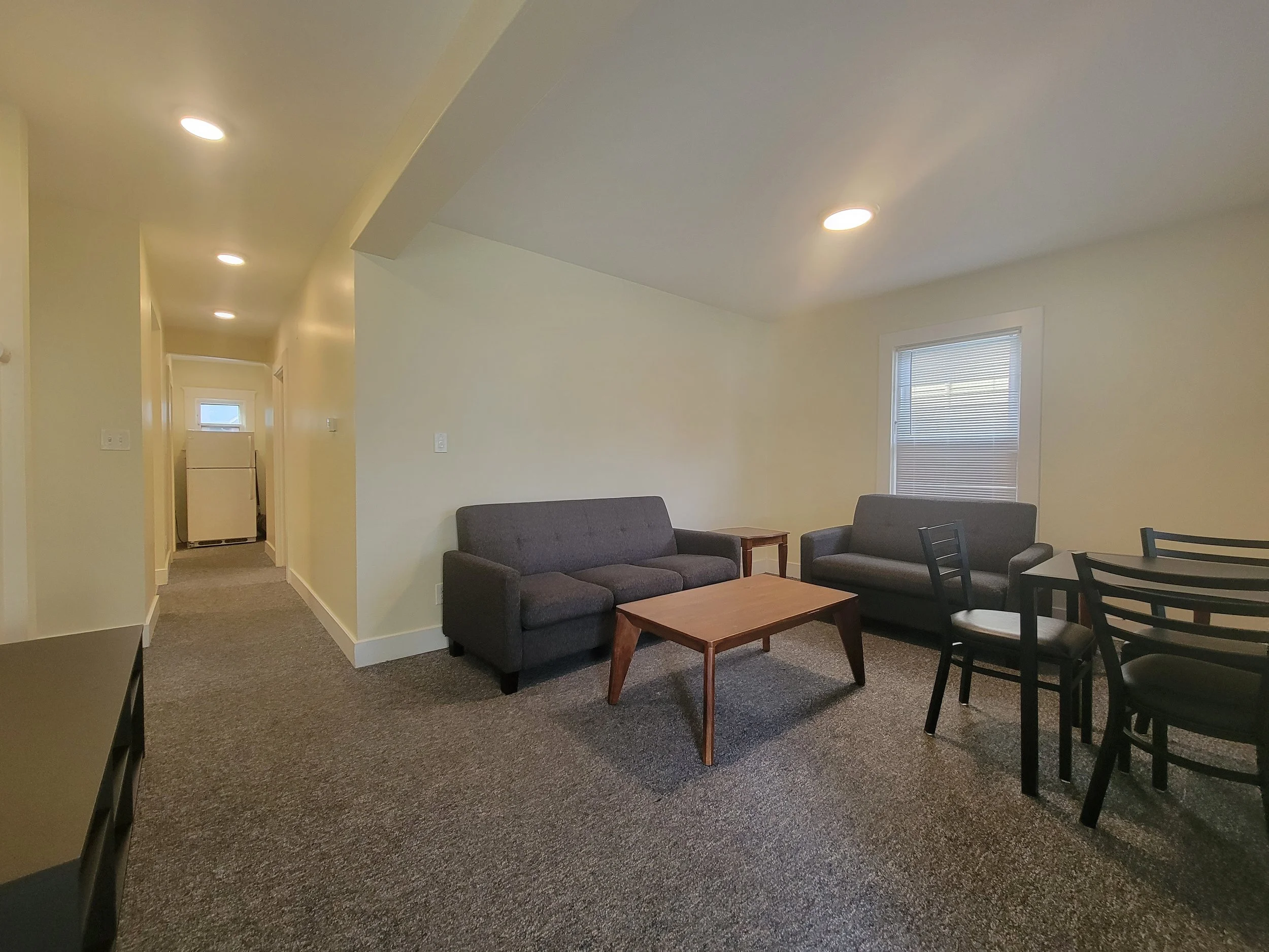 Living room in Ann Arbor for rent with gray sofas, wooden coffee table, small dining set, and hallway leading to a window.