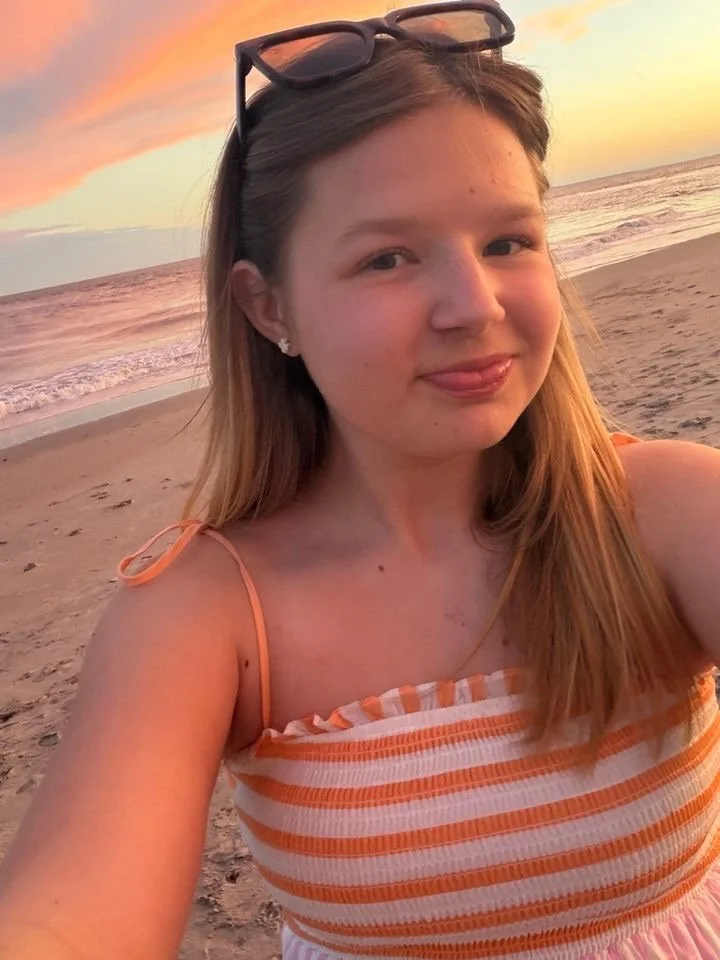 A woman taking a selfie on the beach during sunset, wearing sunglasses on her head and a striped orange and white top.