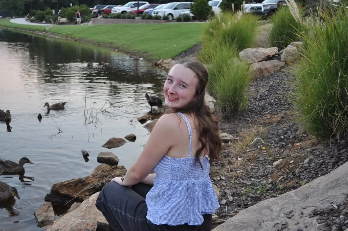 A young woman with long brown hair sitting on rocks by a pond, smiling at the camera with ducks swimming nearby. In the background, there is a grassy area, a park, and parked cars.