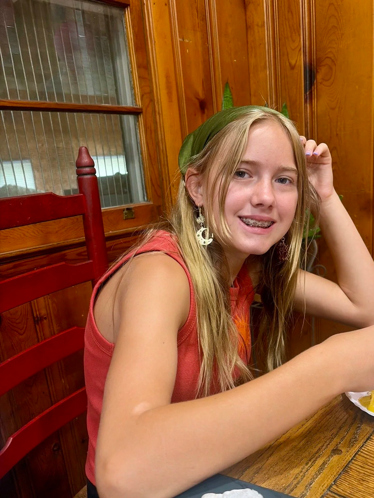 A young woman with long blonde hair, wearing a green headscarf and moon and star earrings, sitting at a wooden table inside a room with wood-paneled walls.
