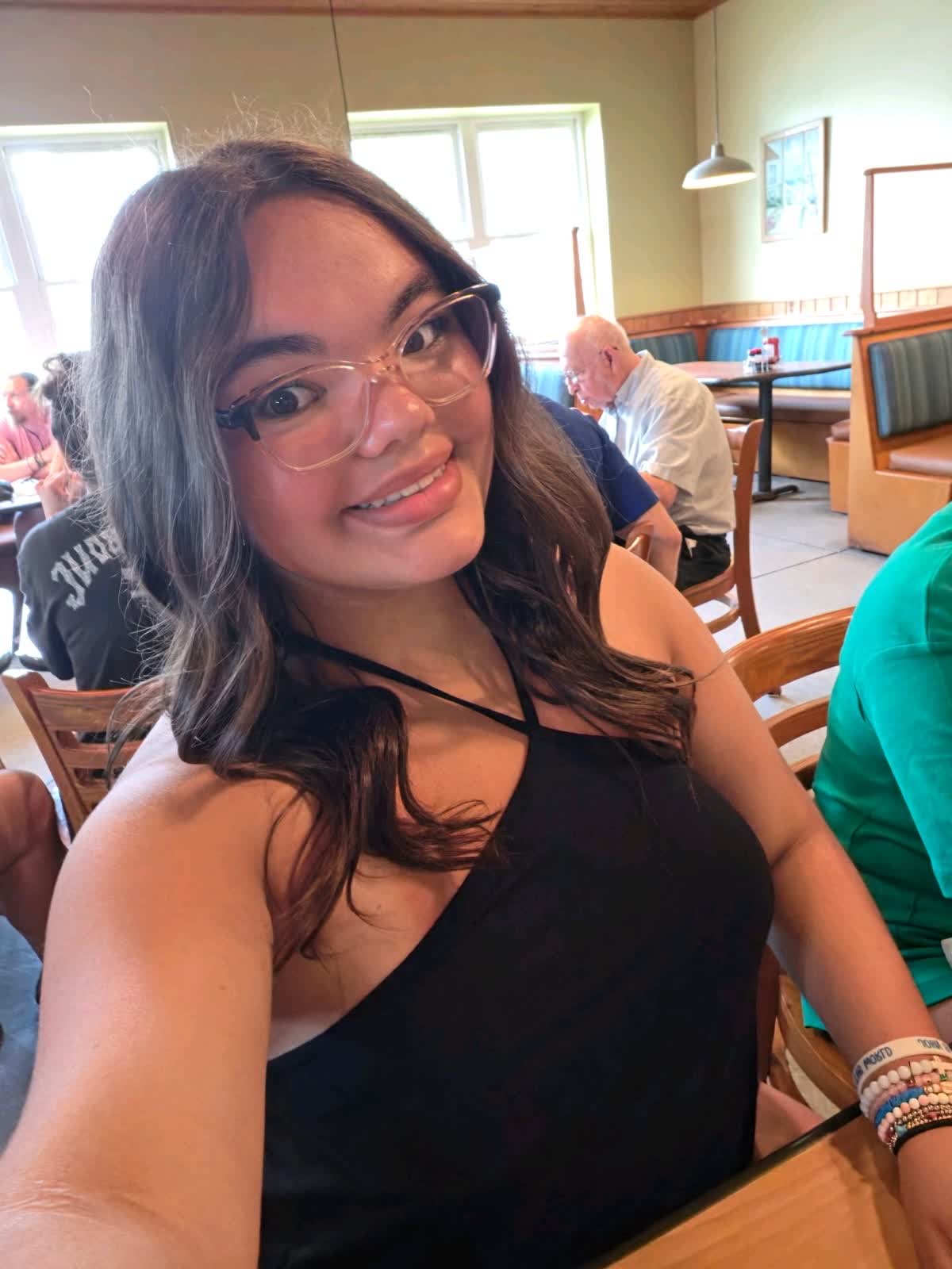 Young woman with glasses and long wavy hair smiling, taking a selfie in a restaurant with other people in the background.