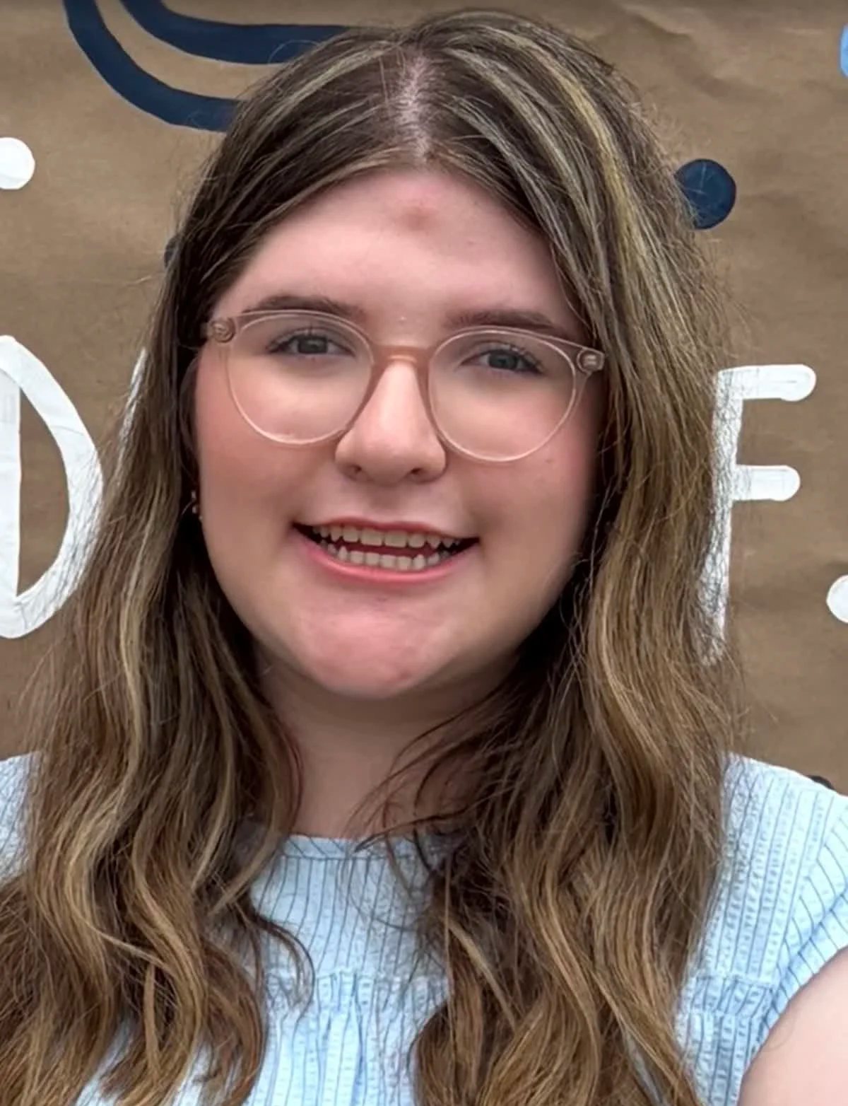A young woman with long wavy brown hair and glasses smiling in front of a brown background with white and blue lettering.