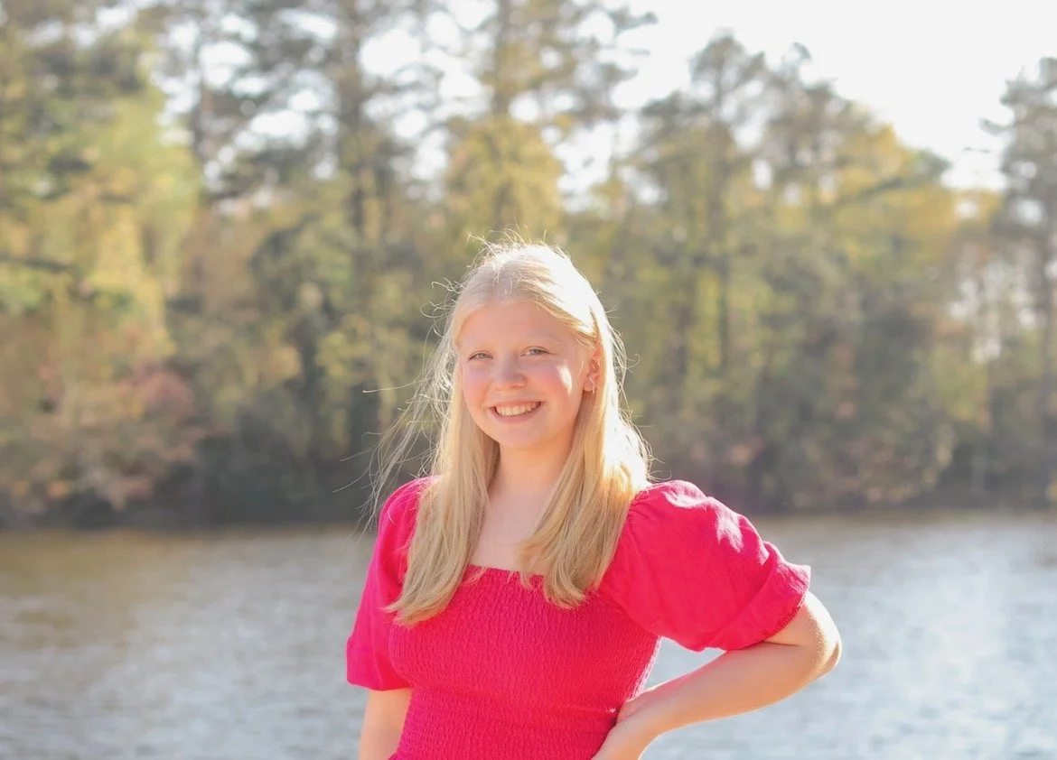 A young girl with long blonde hair smiling outdoors near a body of water with trees in the background, wearing a bright pink top.
