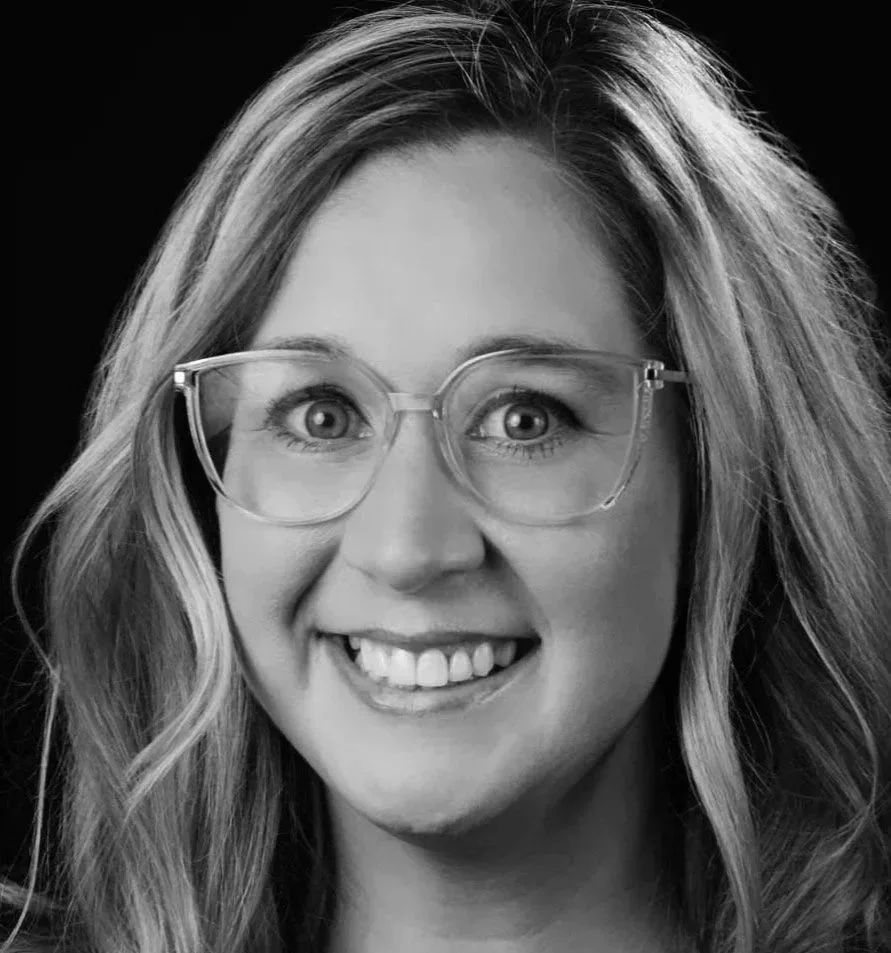 A woman with long wavy hair wearing eyeglasses and smiling against a dark background.