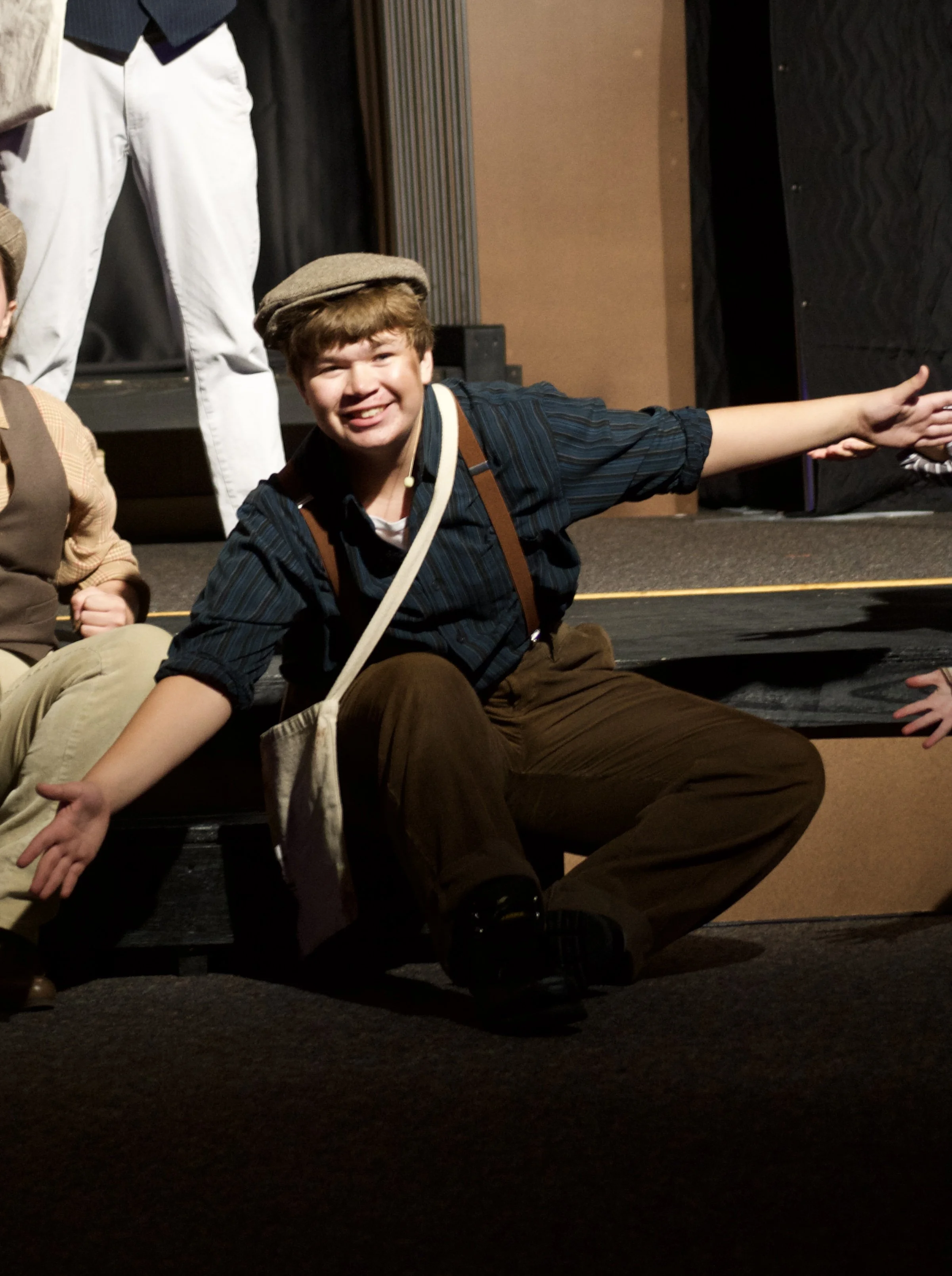 Smiling young man in a flat cap sitting on the stage floor with arms extended, participating in a group activity backstage.
