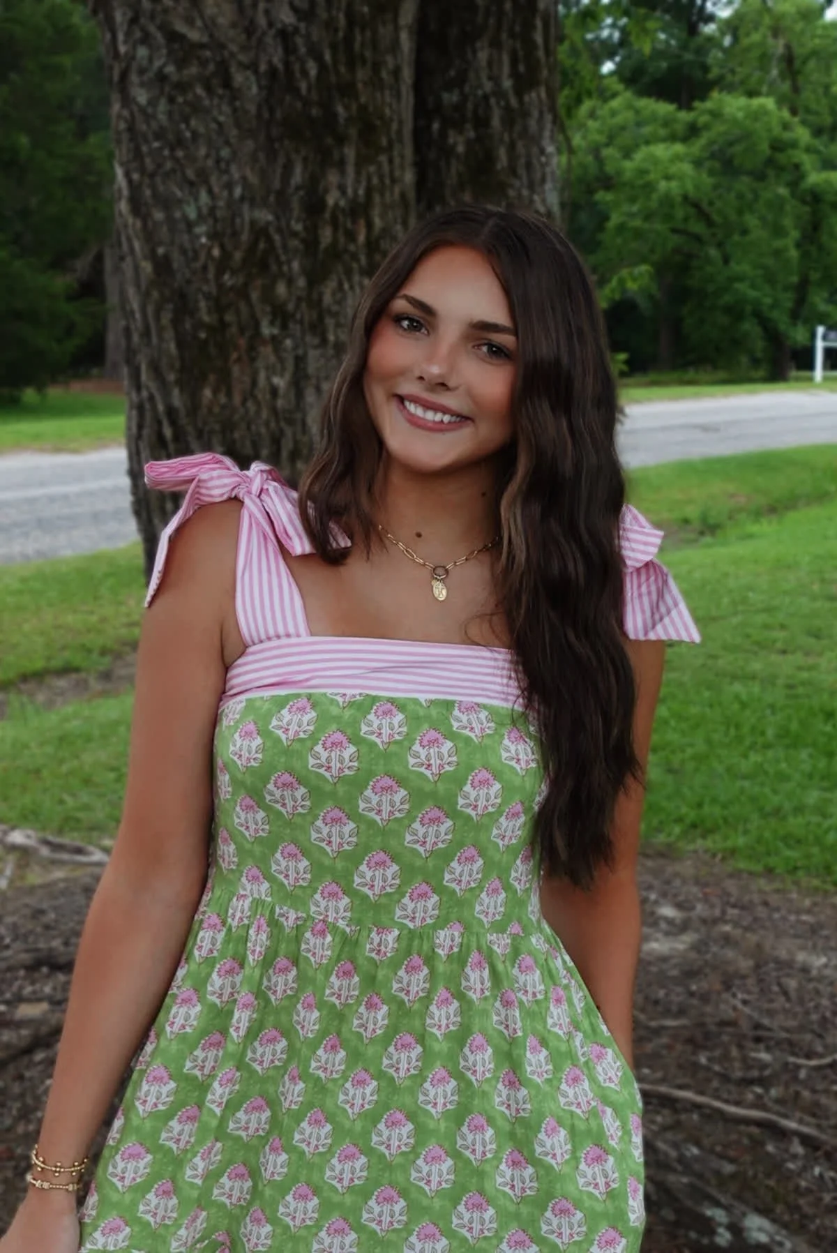 A young woman standing outdoors near a large tree with green trees in the background. She has long, dark wavy hair and is smiling. She is wearing a colorful dress with pink, green, and white patterns, and gold jewelry.