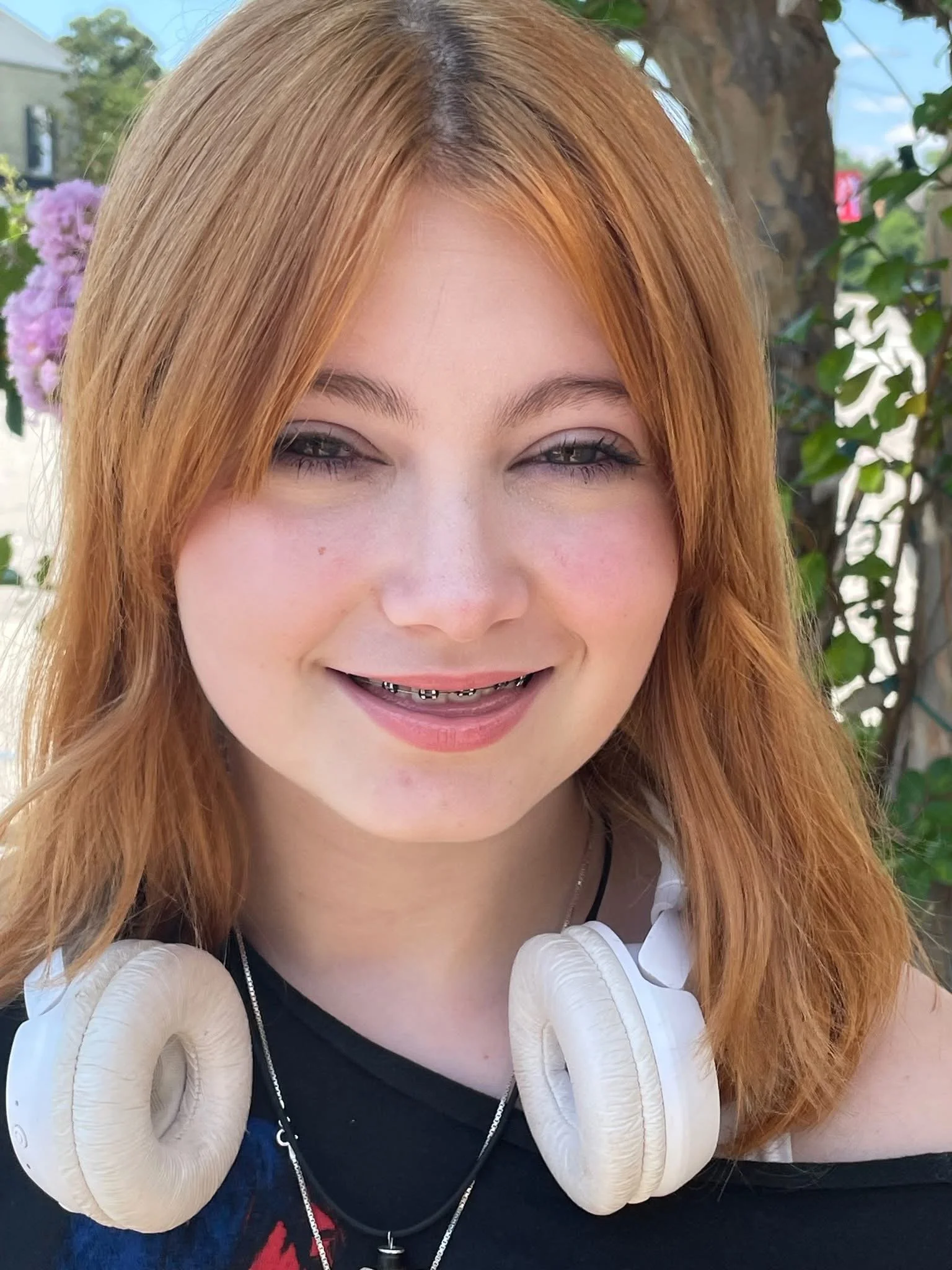 Close-up of a smiling young woman with red hair, wearing a white hoodie and black shirt, with a smiling expression. She has braces and is outdoors with trees and flowers in the background.