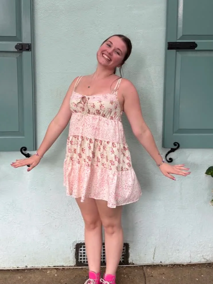 A young woman smiling and posing in front of a light green wall with teal window shutters, wearing a floral pink dress and pink sneakers.