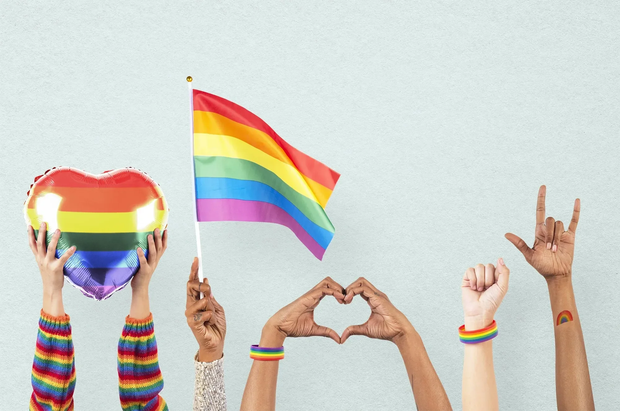 Multiple hands from diverse individuals form various symbols of love, peace, and pride, holding rainbow-colored items including a heart-shaped balloon, a flag, and wristbands, against a light background.