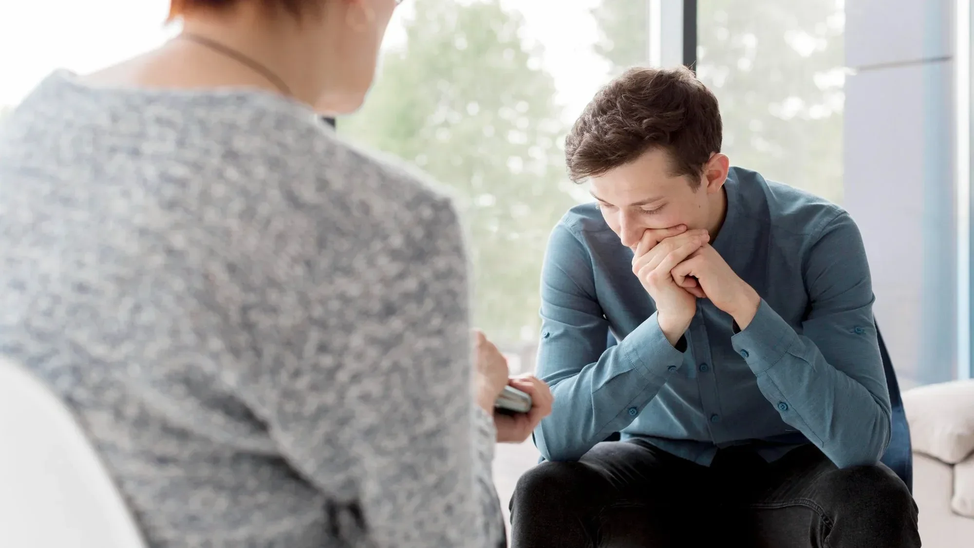 A young man wearing a blue shirt is sitting and praying with his hands clasped near his mouth, seemingly in distress or deep thought. An older woman in the foreground is holding an object, possibly a phone or medication, while sitting in a bright room with large windows showing trees outside.