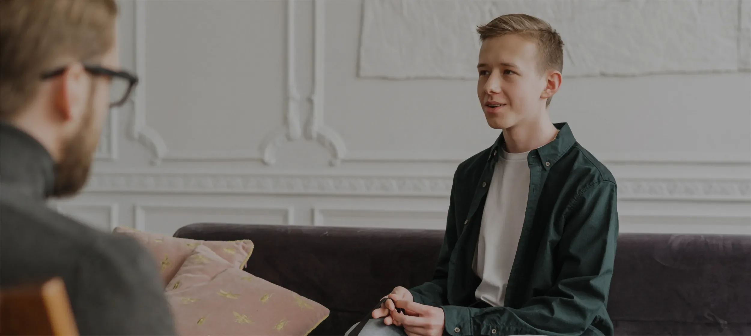 A young man is sitting and speaking with an adult who has glasses and a beard, in a room with white ornate patterns on the wall, a couch with a floral pillow in the foreground.