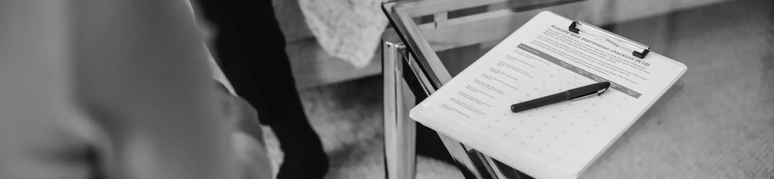A black-and-white photo of a checklist on a clipboard with a pen placed on top, titled 'Anxiety and depression checklist'. The clipboard is resting on a stand or table, and part of a person's arm and a blurred background are visible.