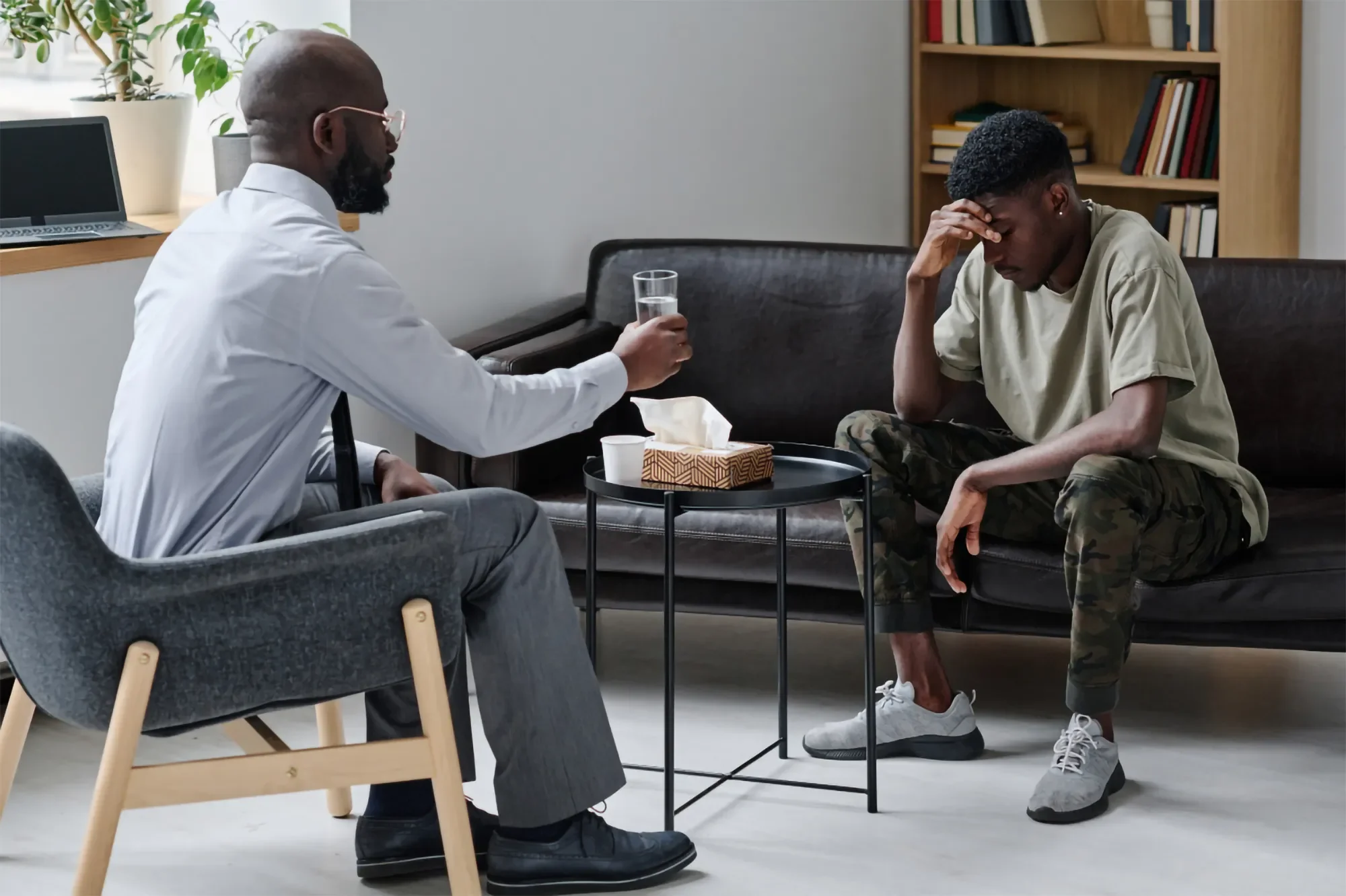 A man in a white shirt and glasses, holding a glass of water, sits on a couch. Another man in a beige t-shirt and camouflage pants, with short black hair, sits on the floor with his hand on his forehead. They are in a room with a bookshelf, a window with plants, and a small table with tissues and a cup.