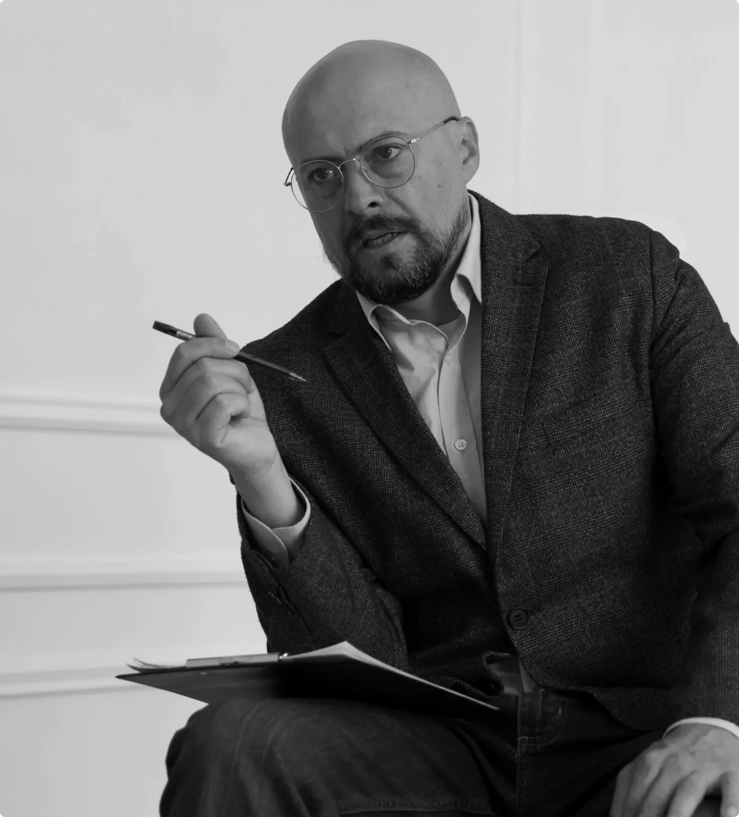 Black and white photo of a bald man with glasses seated at a table, holding a pen and looking to his left, with folder or notebook on his lap.