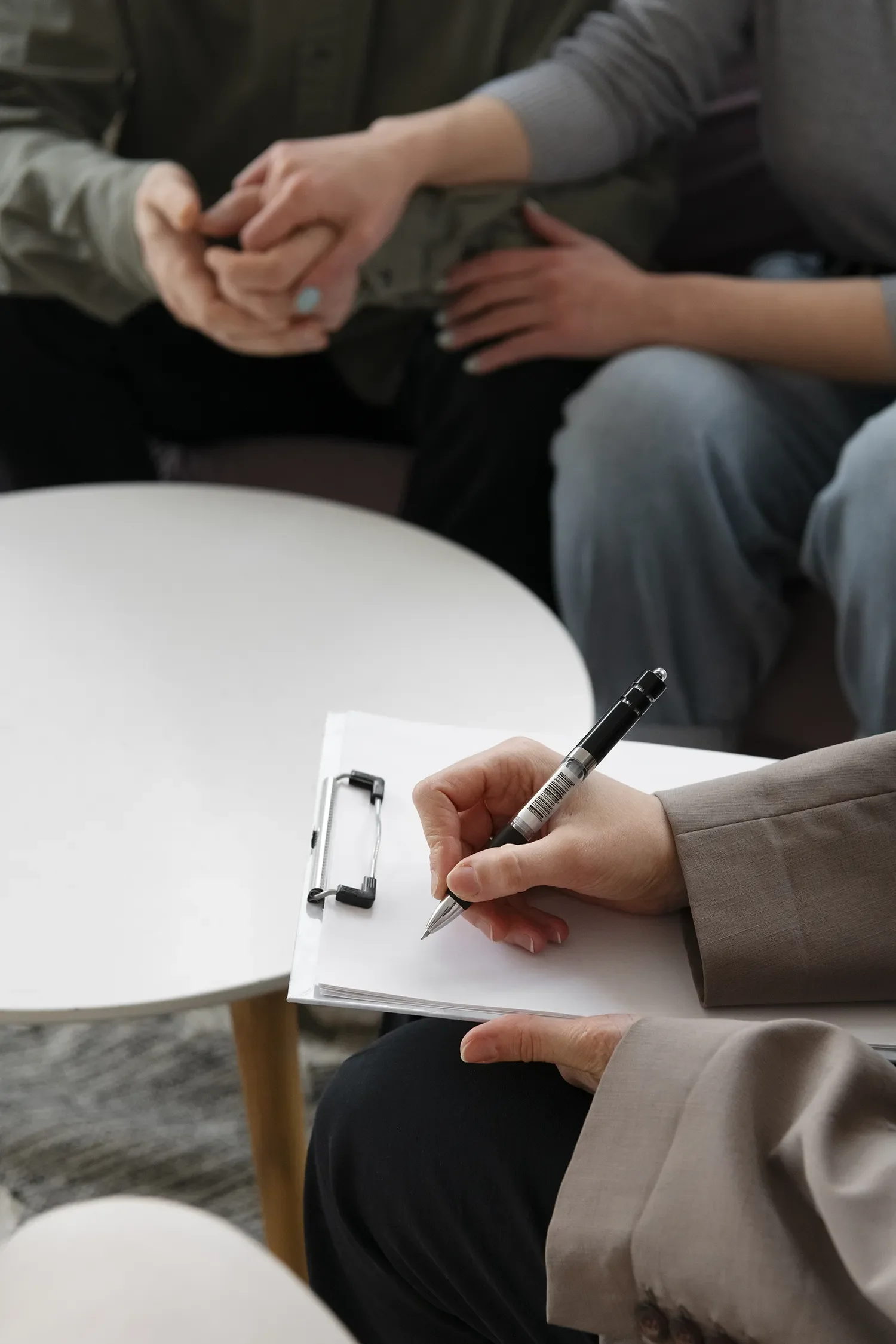 A person in a beige blazer is writing on a clipboard with a black pen, sitting at a white table. Another person, wearing a gray top, is holding hands with someone else in the background.