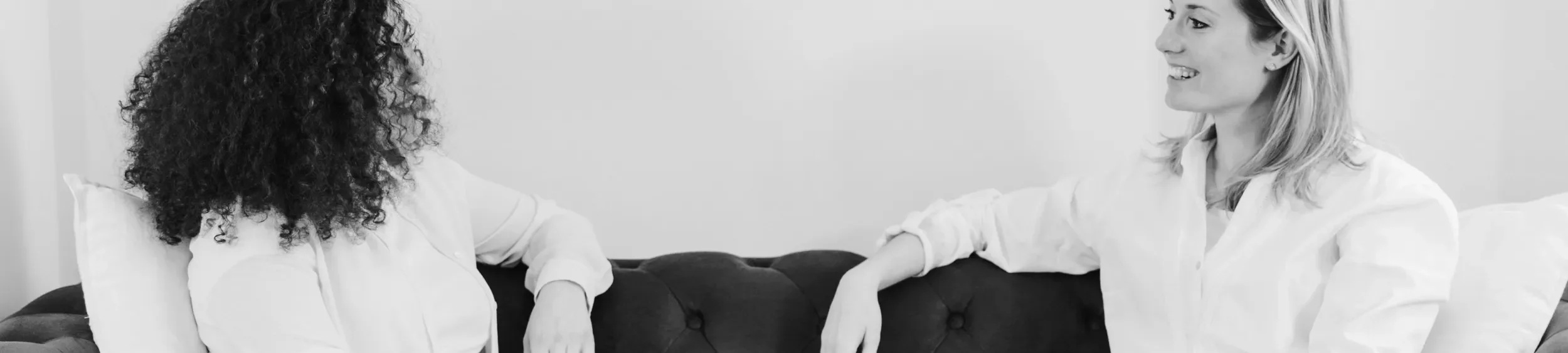 Two women sitting on a couch engaged in conversation, smiling, one with curly hair and the other with straight hair, both wearing white shirts.