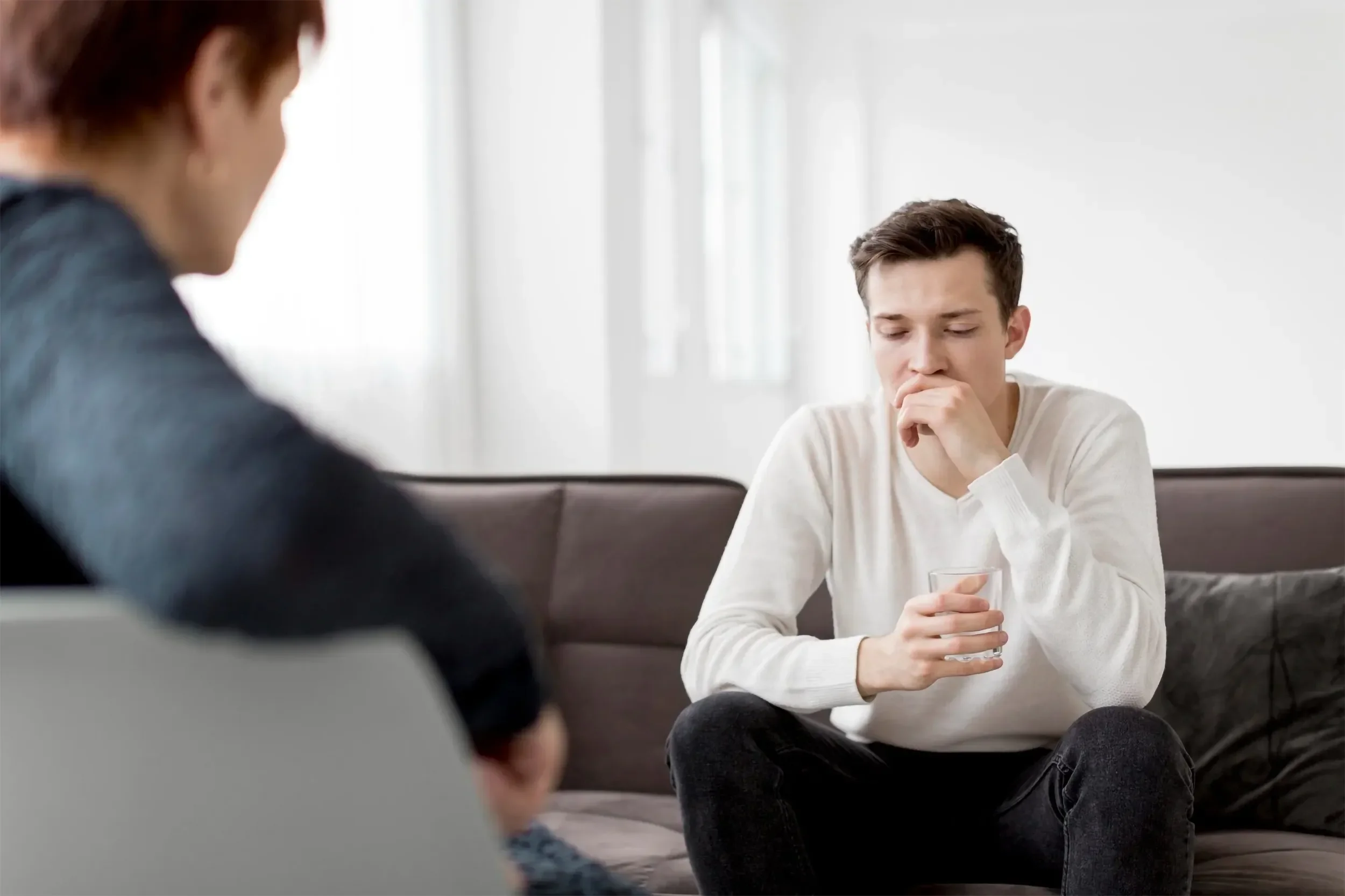 A young man is sitting on a sofa, holding a glass of water, and appearing thoughtful. Two people are partially visible in the foreground, engaged in conversation. The setting is a bright, minimalistic room with large windows.