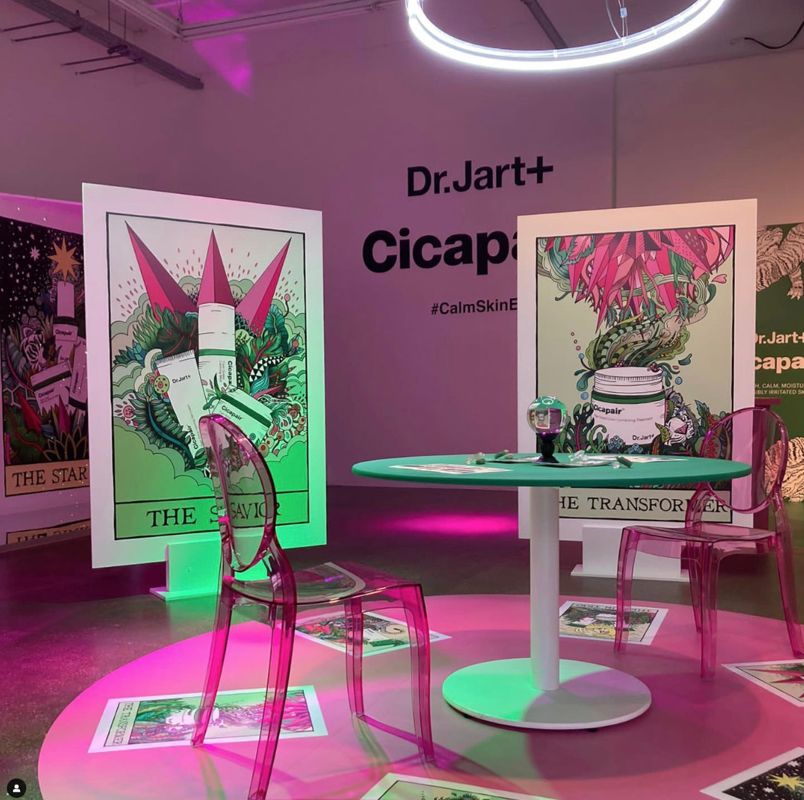 An indoor promotional display with colorful illustrations of skincare products, transparent pink chairs, and a round green table. There are large posters with artistic designs and text related to Dr. Jart+ and Cicapair products, with purple and pink 