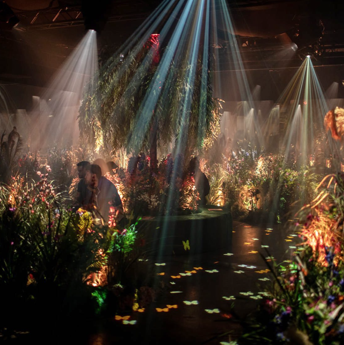 Indoor event space decorated with lush greenery and colorful flowers, illuminated by spotlights creating a dreamy, enchanting atmosphere.