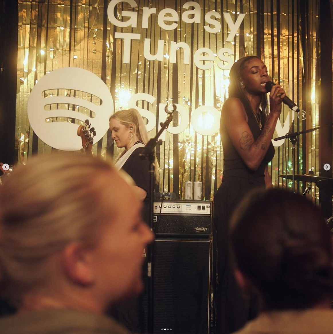 Two women performing on stage at Greasy Tunes, with a backdrop of golden metallic streamers, a large Spotify logo, and the words "Greasy Tunes" on a sign.