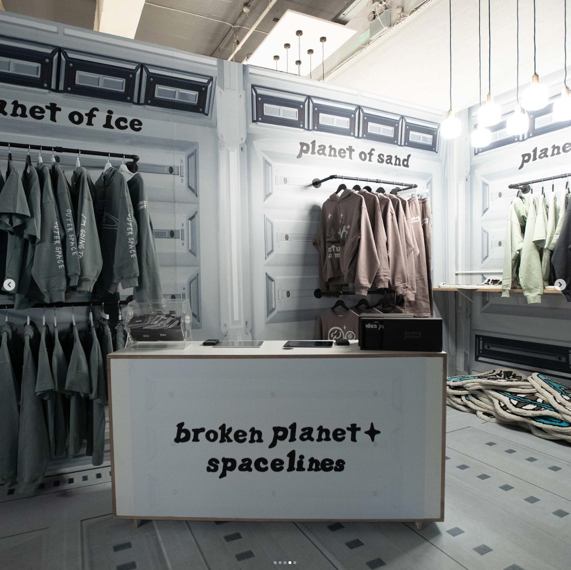 Retail clothing display with hoodies on racks, a sign reading "broken planet + spacelines," and background signs "planet of ice" and "planet of sand" in a sci-fi themed store.