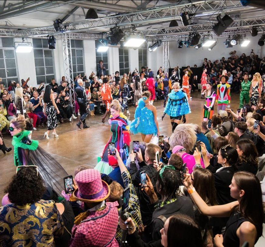 Fashion show with models on runway showcasing colorful outfits, audience taking photos, and bright lighting.