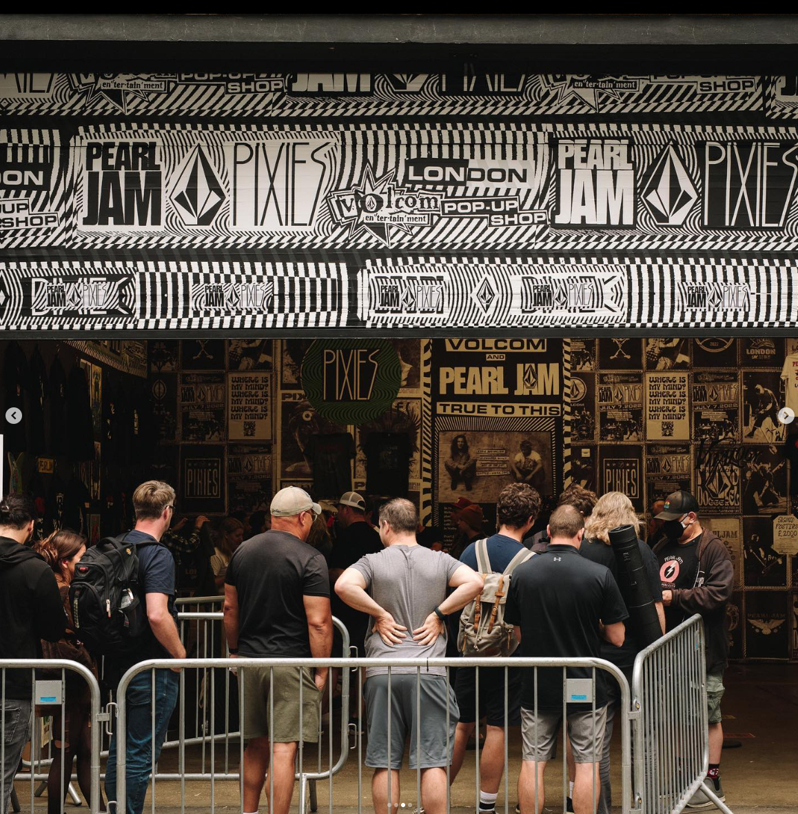 People gathered inside a poster-filled room at a Pearl Jam and Pixies concert or event. The posters on the wall feature band logos and concert information, and there is a skyline illustration with the words "London" and "Pop-Up Shop". The crowd is st