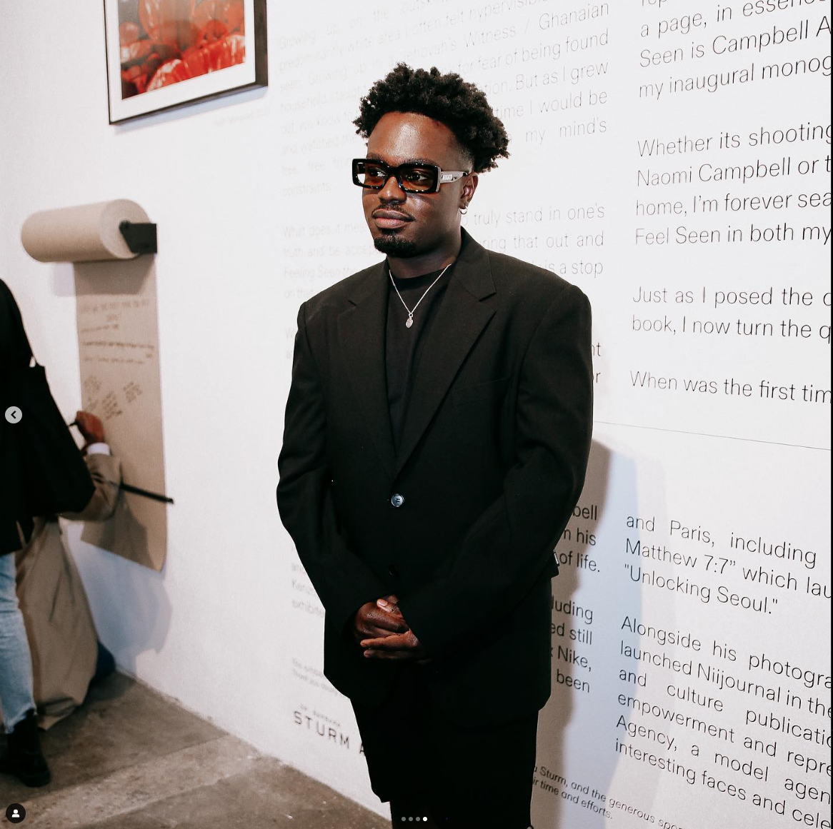 A man in a black suit and glasses standing in front of a white wall with printed text, with a framed picture and a person writing on a large pad in the background.