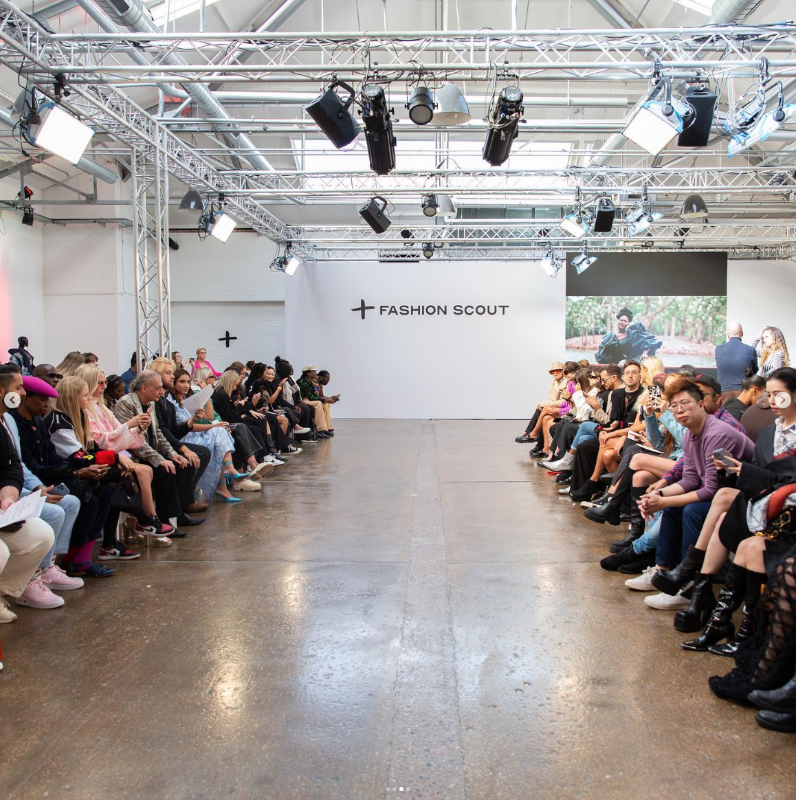 Fashion show runway with seated audience on both sides, a large white wall with '+FASHION SCOUT' logo, set in an industrial-style venue with lighting equipment overhead, and a large screen displaying a fashion image in the background.