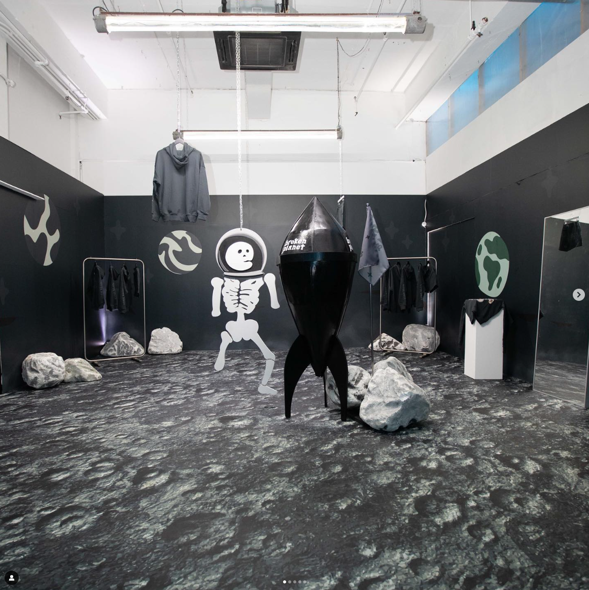 Room with lunar surface floor, rocks, black walls, and space-themed decorations including a skeleton cartoon figure, an astronaut helmet, and a rocket model labeled 'Broken Planet'. Clothes hanging on racks, a hoodie hanging from the ceiling, and ind