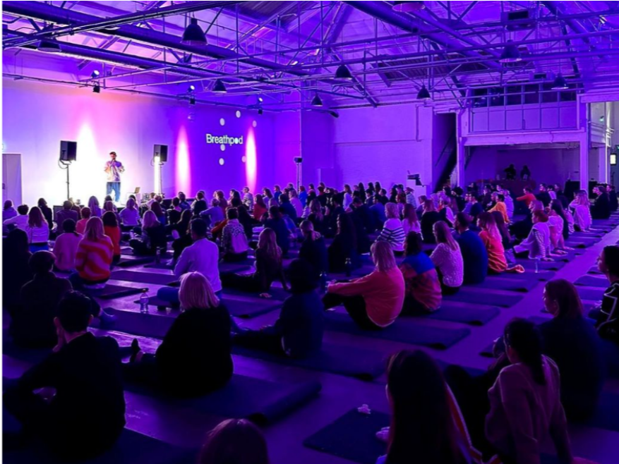 Large group of people sitting on yoga mats in a spacious industrial-style event hall, attending a presentation or workshop with purple lighting and a speaker on stage.