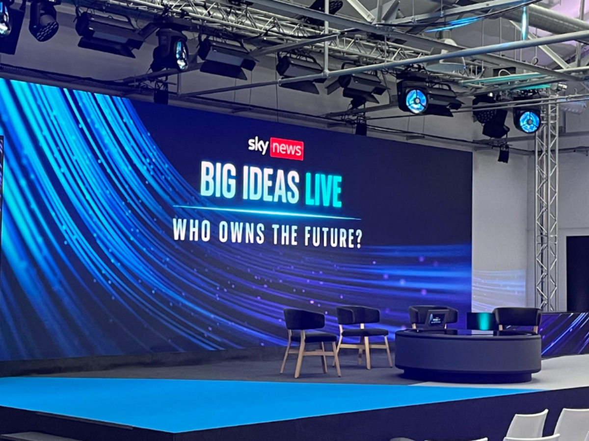 Stage setup for a Sky News event titled 'Big Ideas Live' with the question 'Who owns the future?' on a large digital screen. The stage has three chairs, a round table with a laptop, and is illuminated with blue lighting and overhead stage lights.